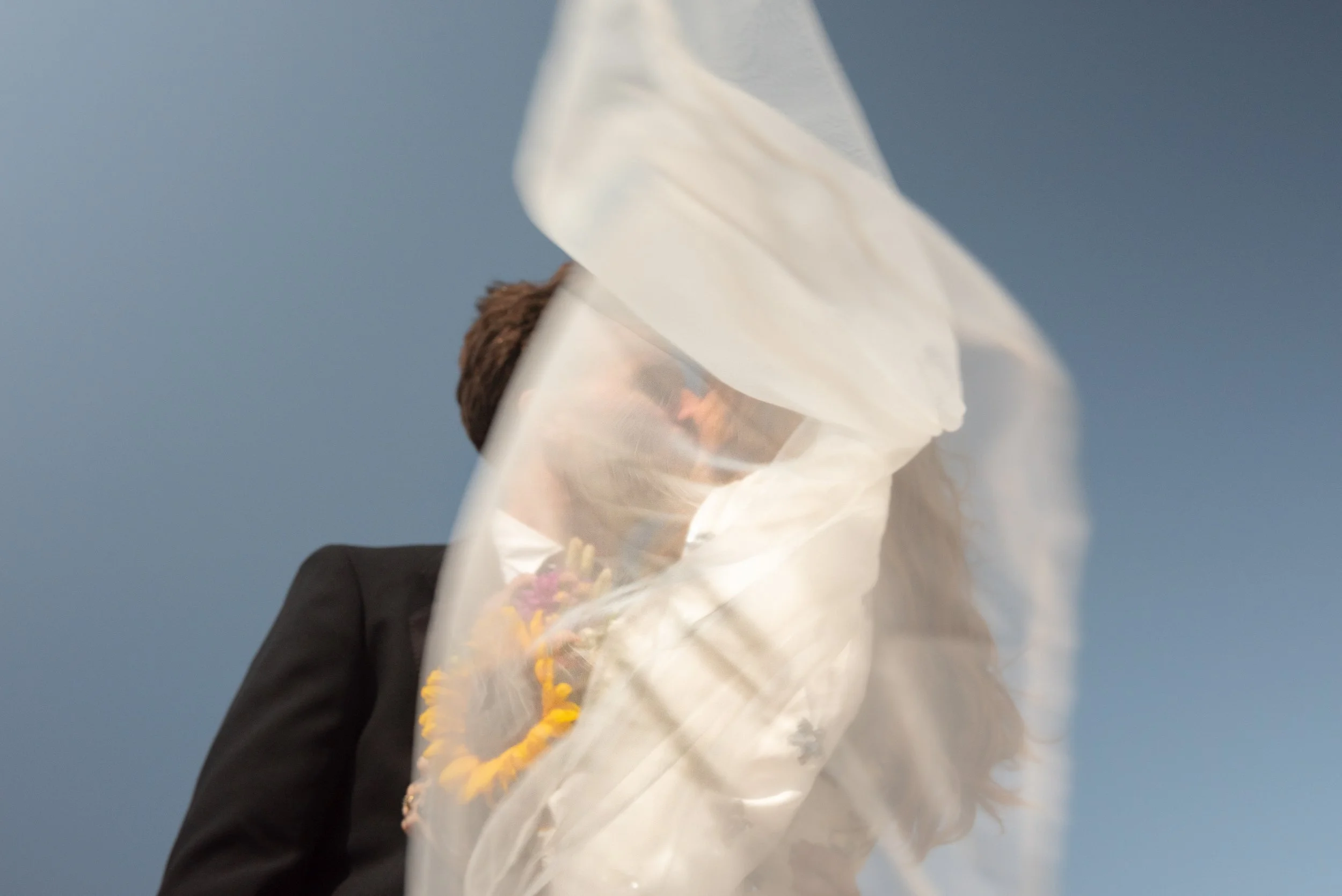 A person with brown hair wearing a black suit holding a bouquet of flowers, partially obscured by a large piece of white fabric or paper.
