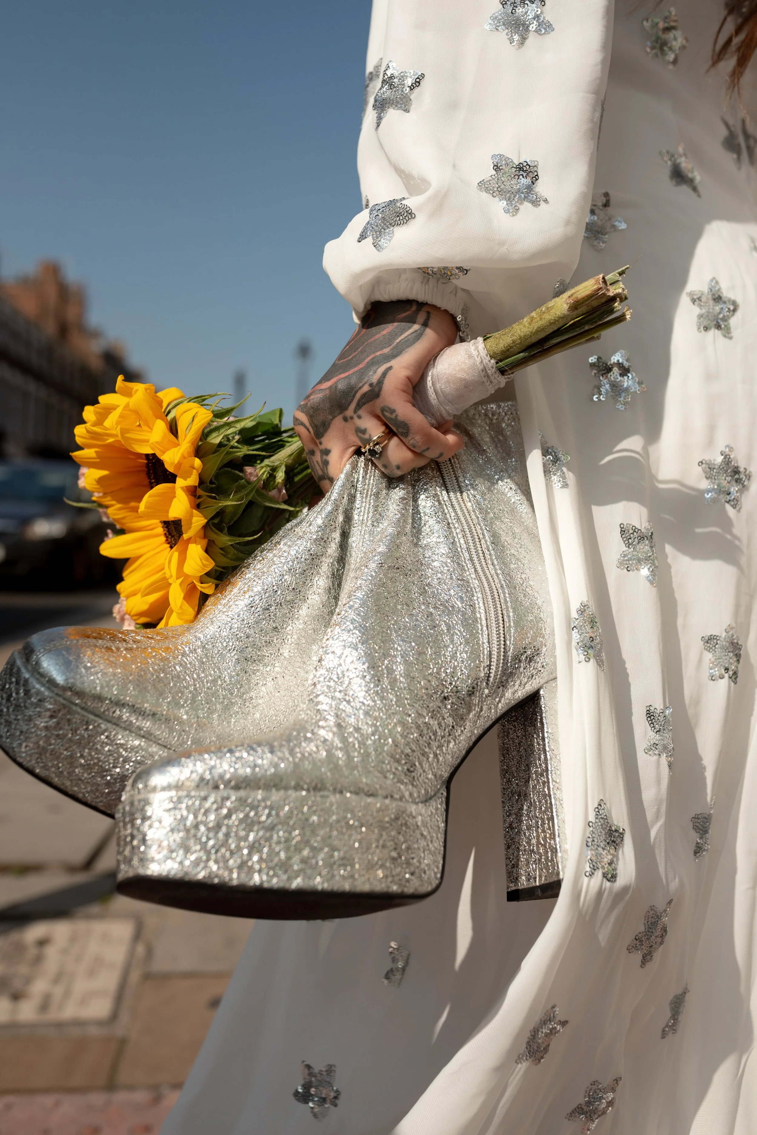 A person holding a bouquet of yellow sunflowers while balancing a shiny silver high-heeled boot against a white fabric adorned with silver sequin butterfly embellishments.