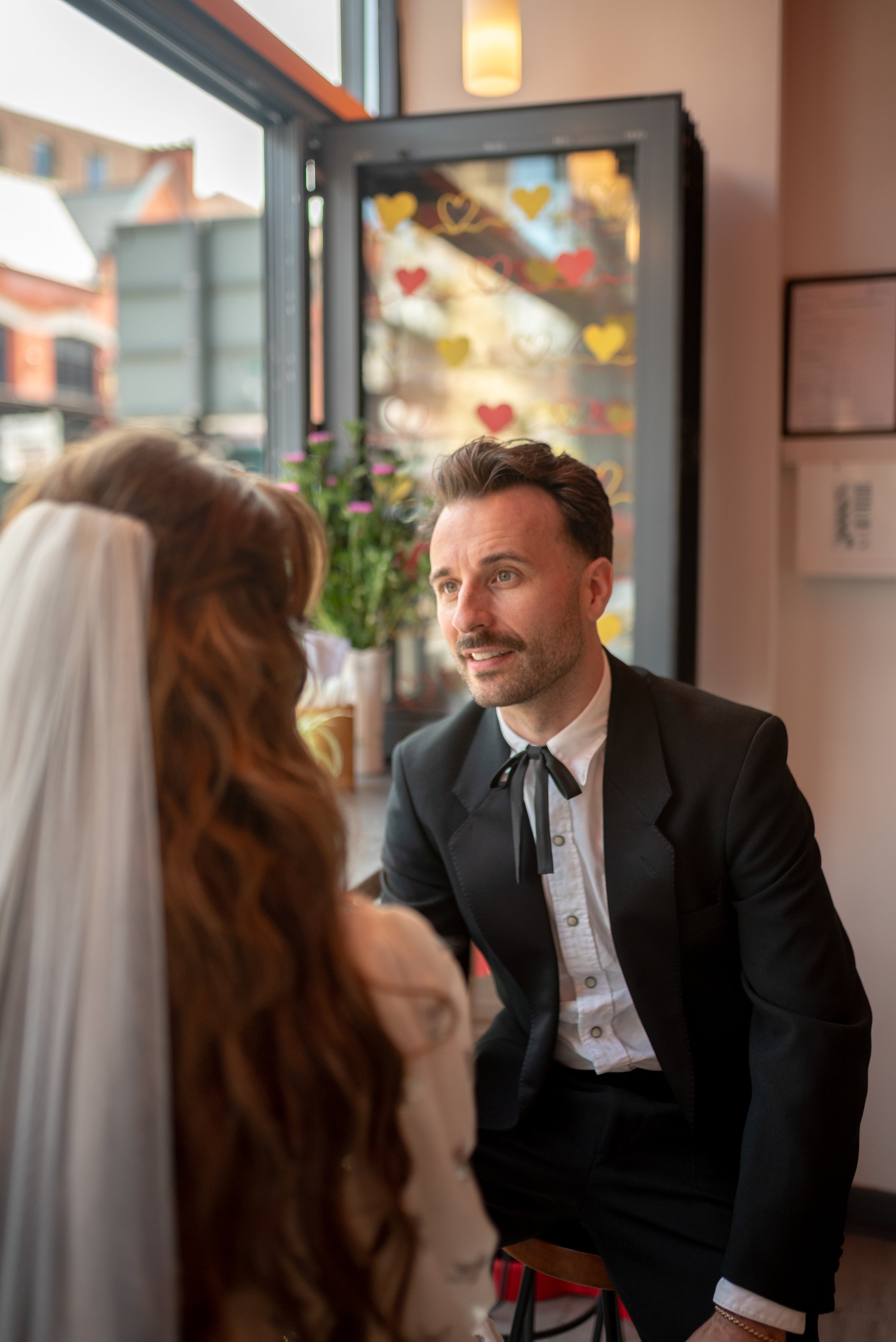 A groom in a black tuxedo speaking with a bride with long red hair and a white veil in a well-lit cafe with heart decorations on the window.