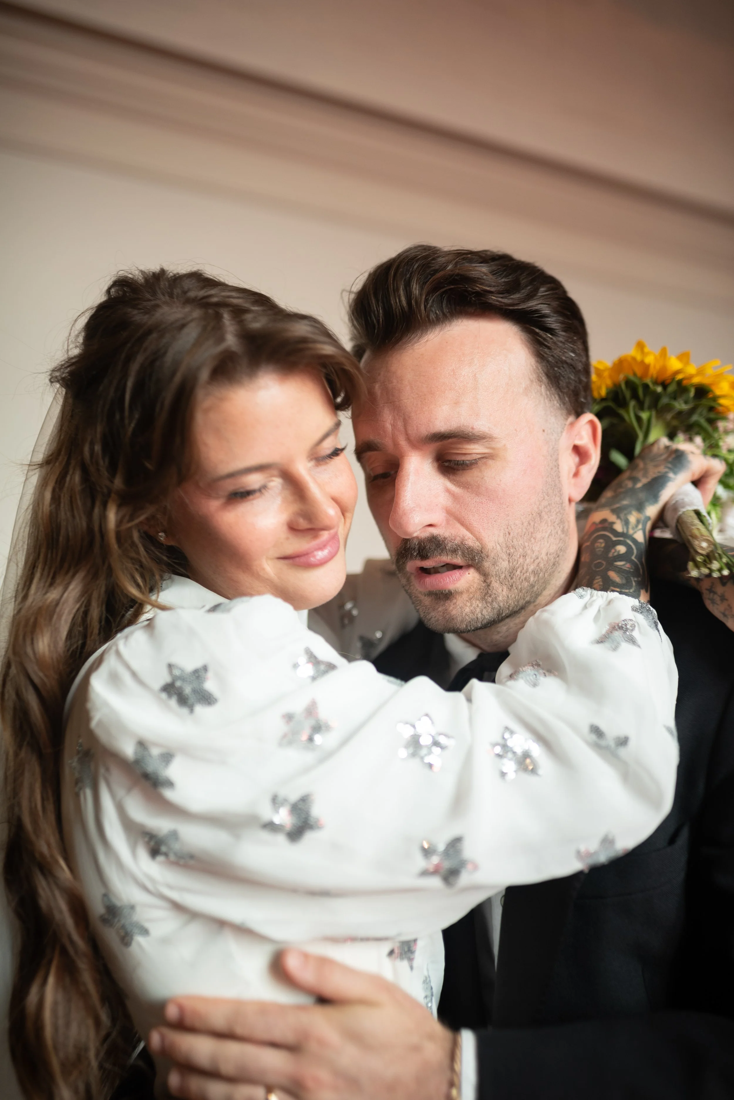 A woman and a man embrace closely, with the woman holding a bouquet of sunflowers behind the man's shoulder. They appear to be happy and intimate.