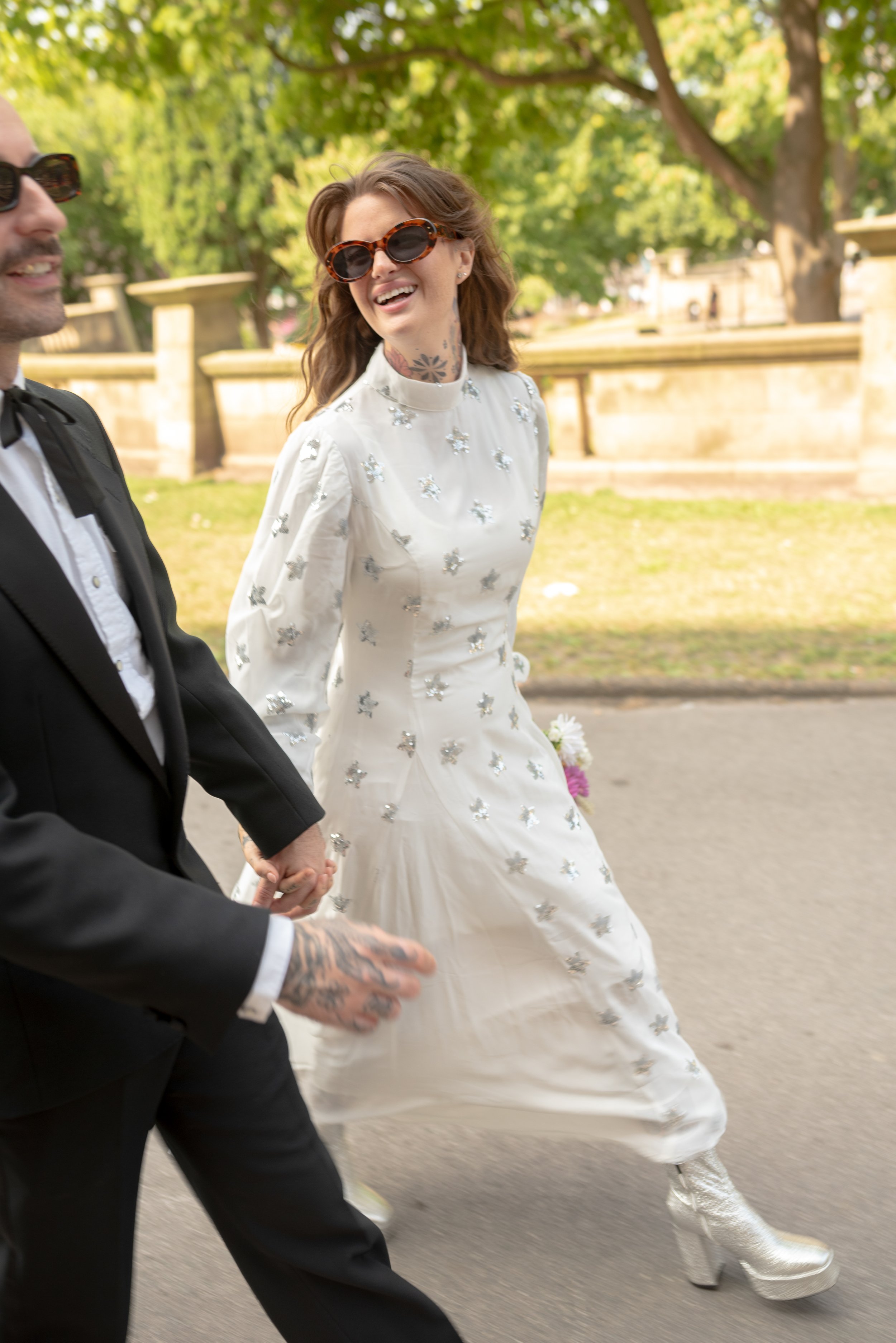 A woman wearing large sunglasses and a white dress with silver embellishments, holding hands with a man in a black tuxedo with tattoos on his hand, walking outdoors in a park with green trees and stone fences in the background.