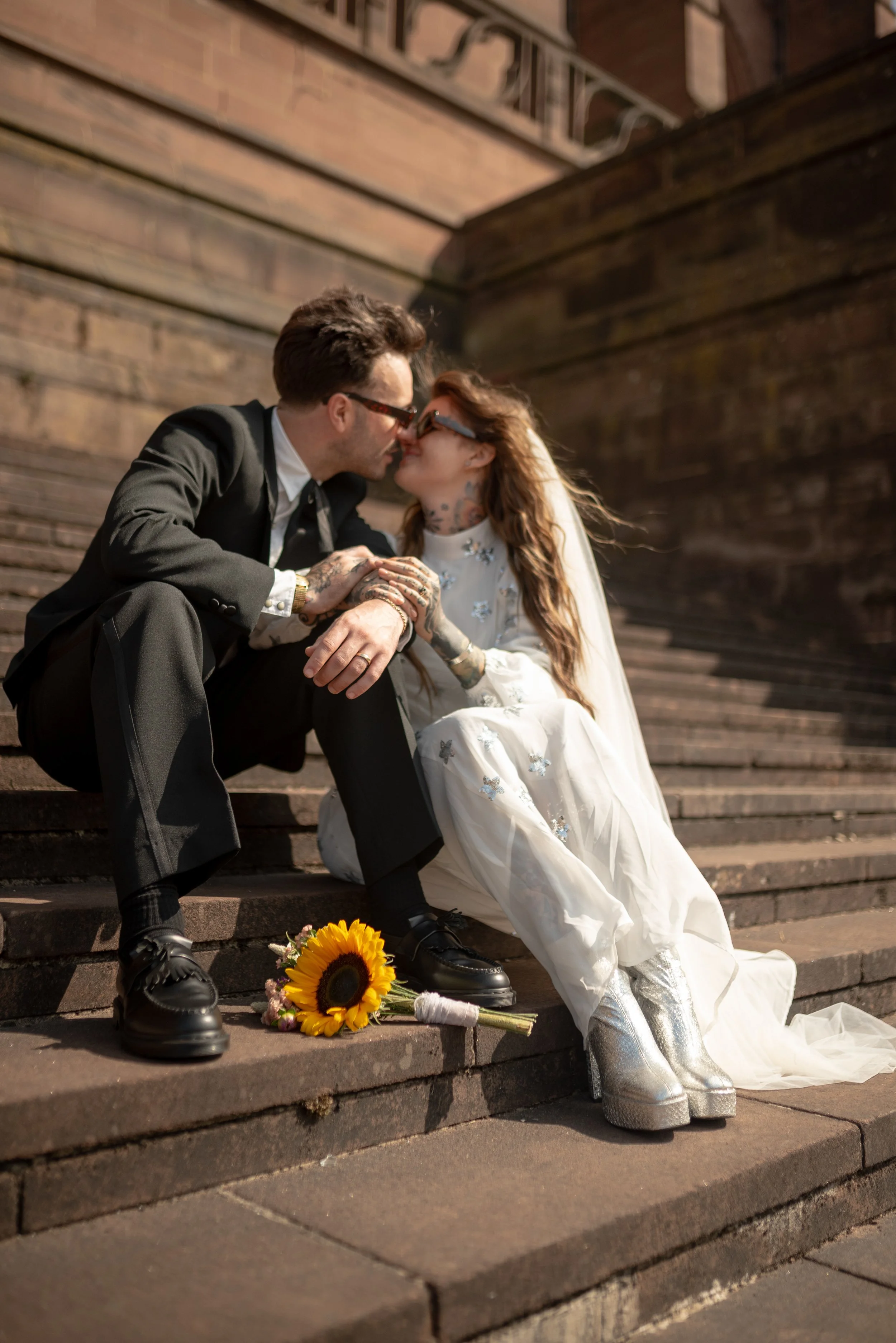 A couple sitting on outdoor stairs, dressed in wedding attire, kissing. The bride is wearing a white gown with floral embroidery and silver platform boots, and the groom is in a black suit and sunglasses. A bouquet of sunflowers rests on the stairs b