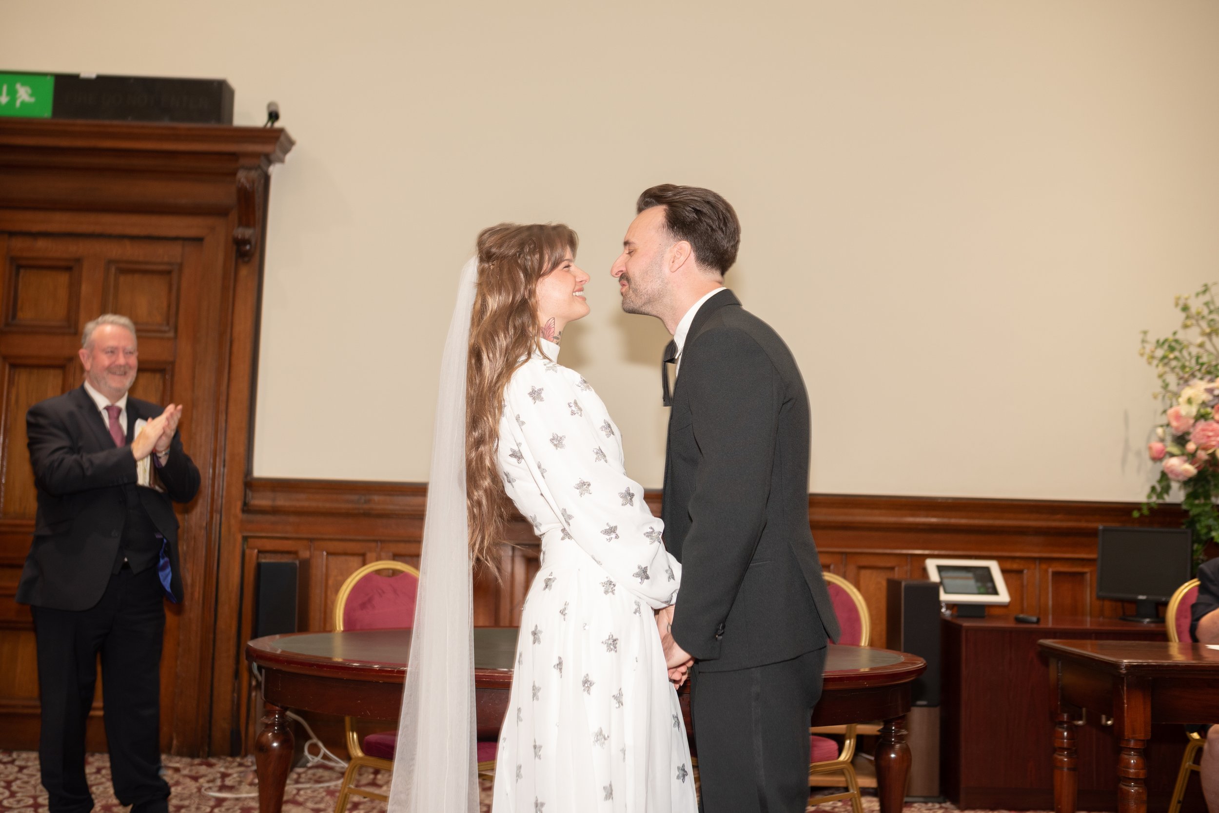 A bride and groom holding hands and facing each other during their wedding ceremony in a wood-paneled room with a man clapping in the background.