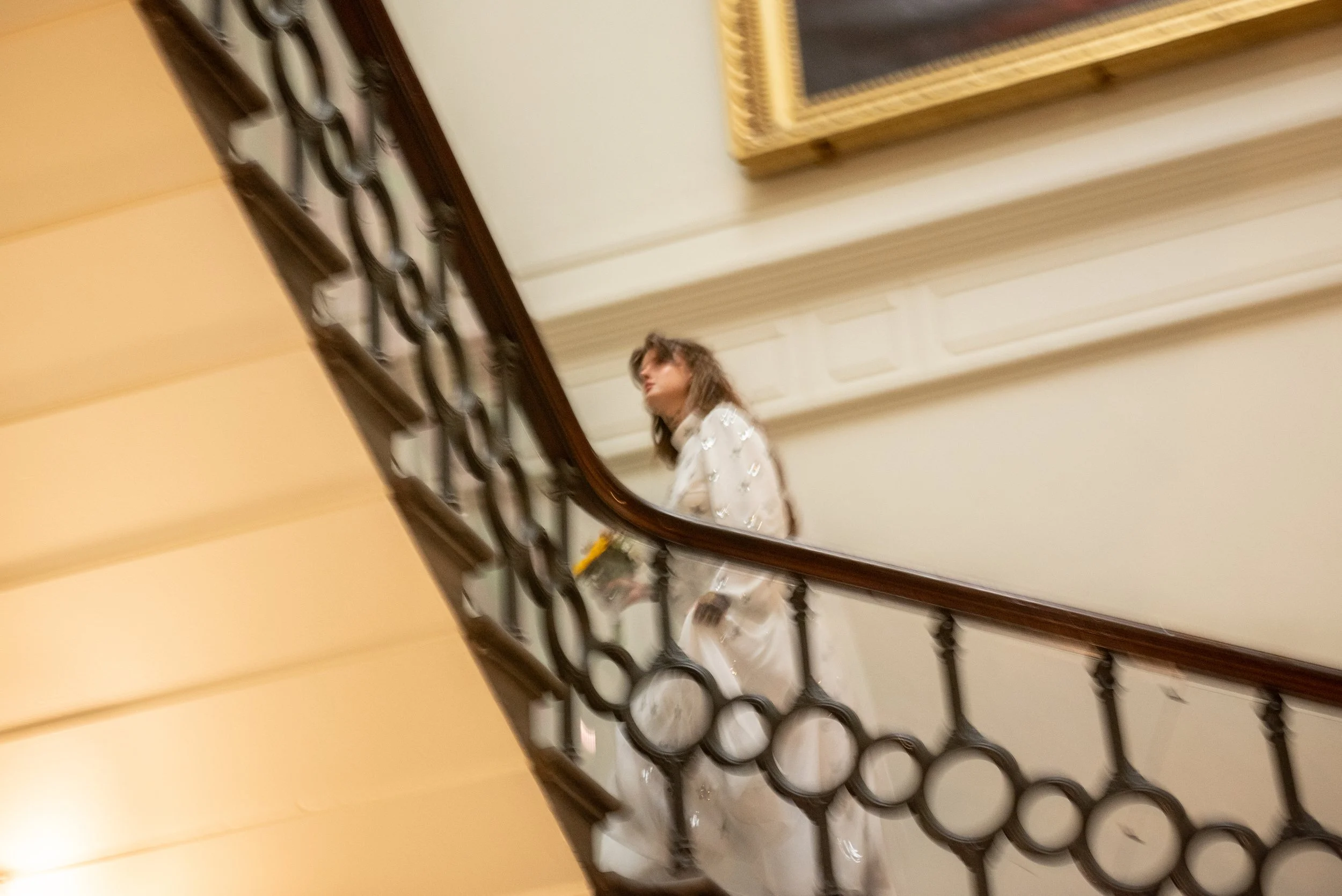 A woman in a white dress carrying a bouquet of flowers walking down a staircase with a decorative iron railing.