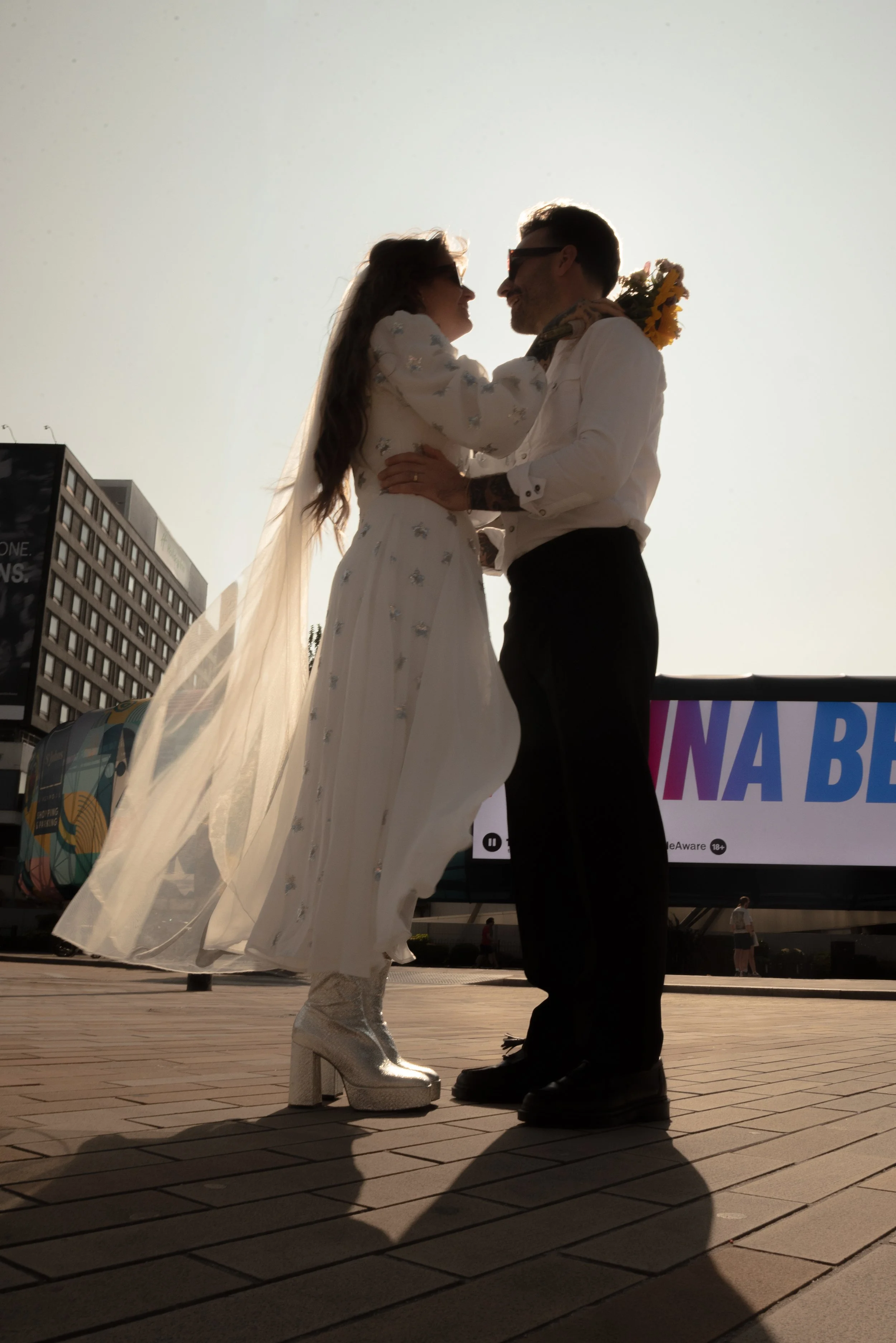 A couple dressed in wedding attire sharing a moment outdoors with a bright sky background, a billboard, and city buildings in the distance.