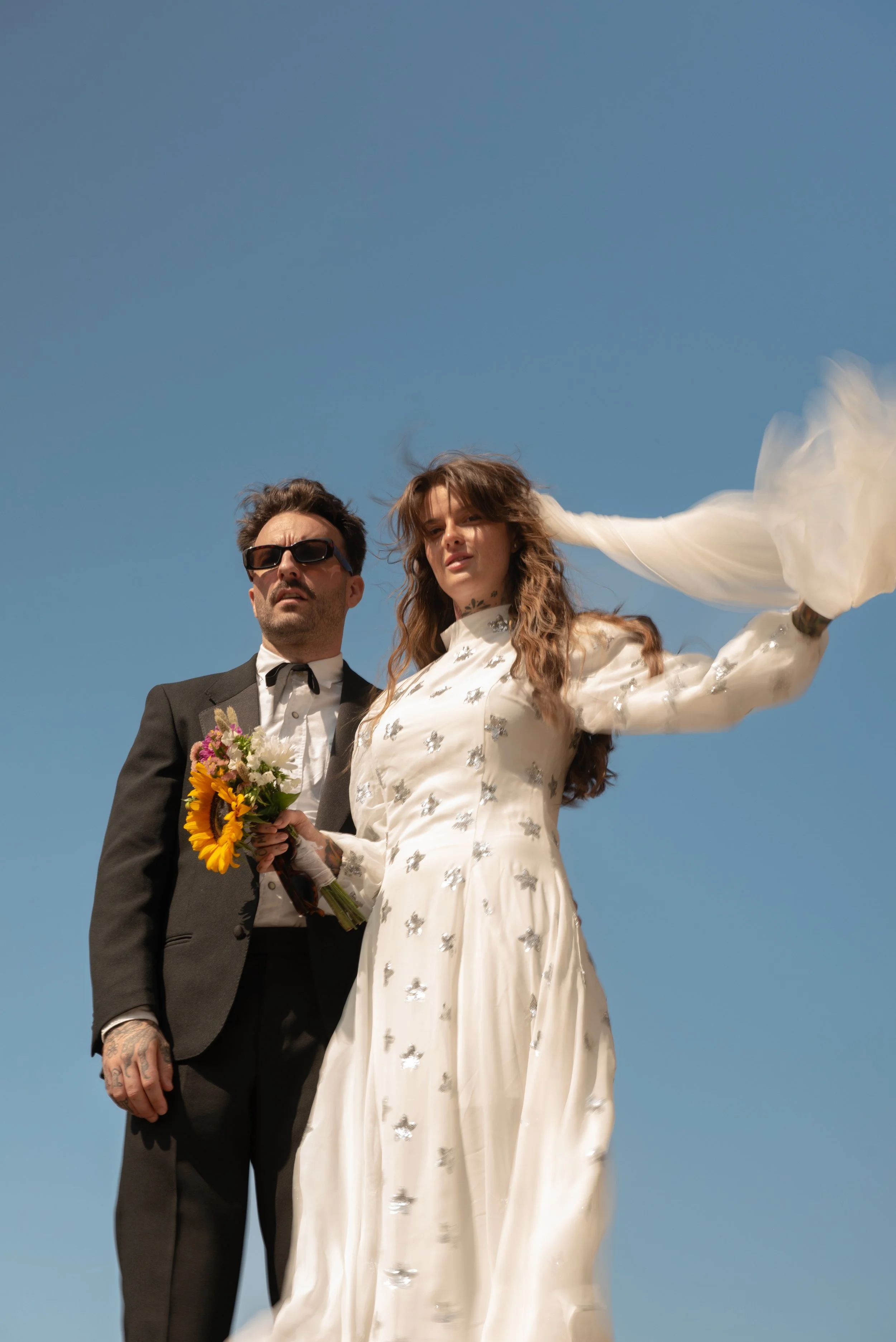 A man in a suit and sunglasses and a woman in a white dress holding a bouquet of flowers stand against a clear blue sky, looking into the distance.