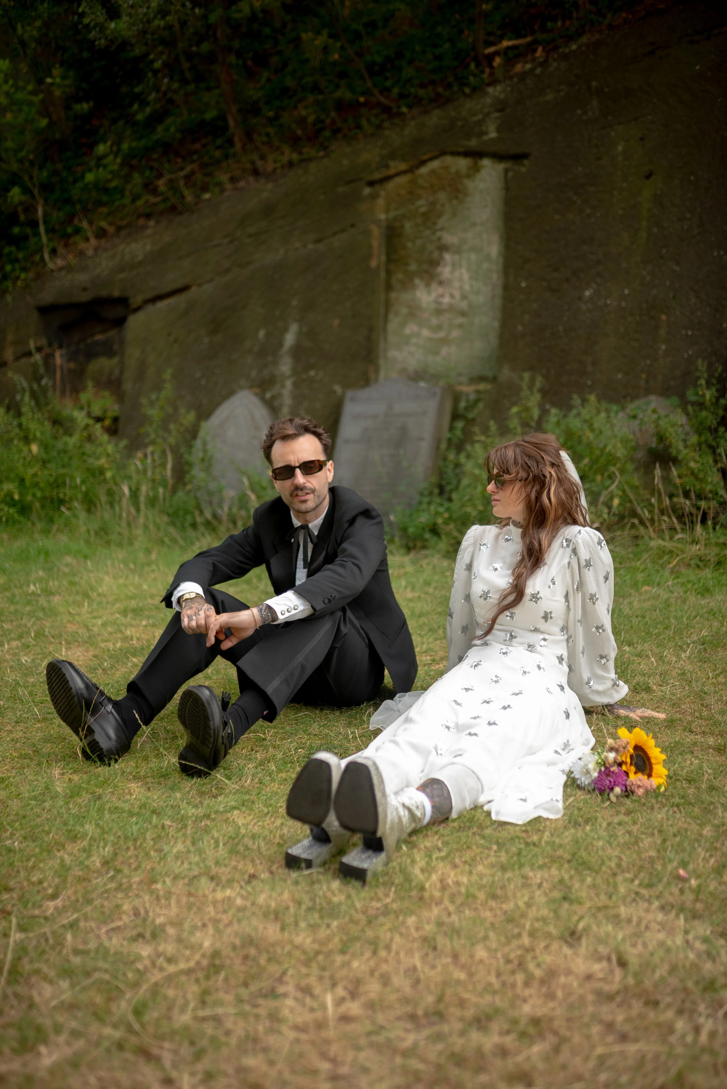 A man and woman sitting on the grass in front of a gravestone, with the woman wearing roller skates and a dress, and the man in a black suit, sunglasses, and tattoos, in a grassy outdoor setting.