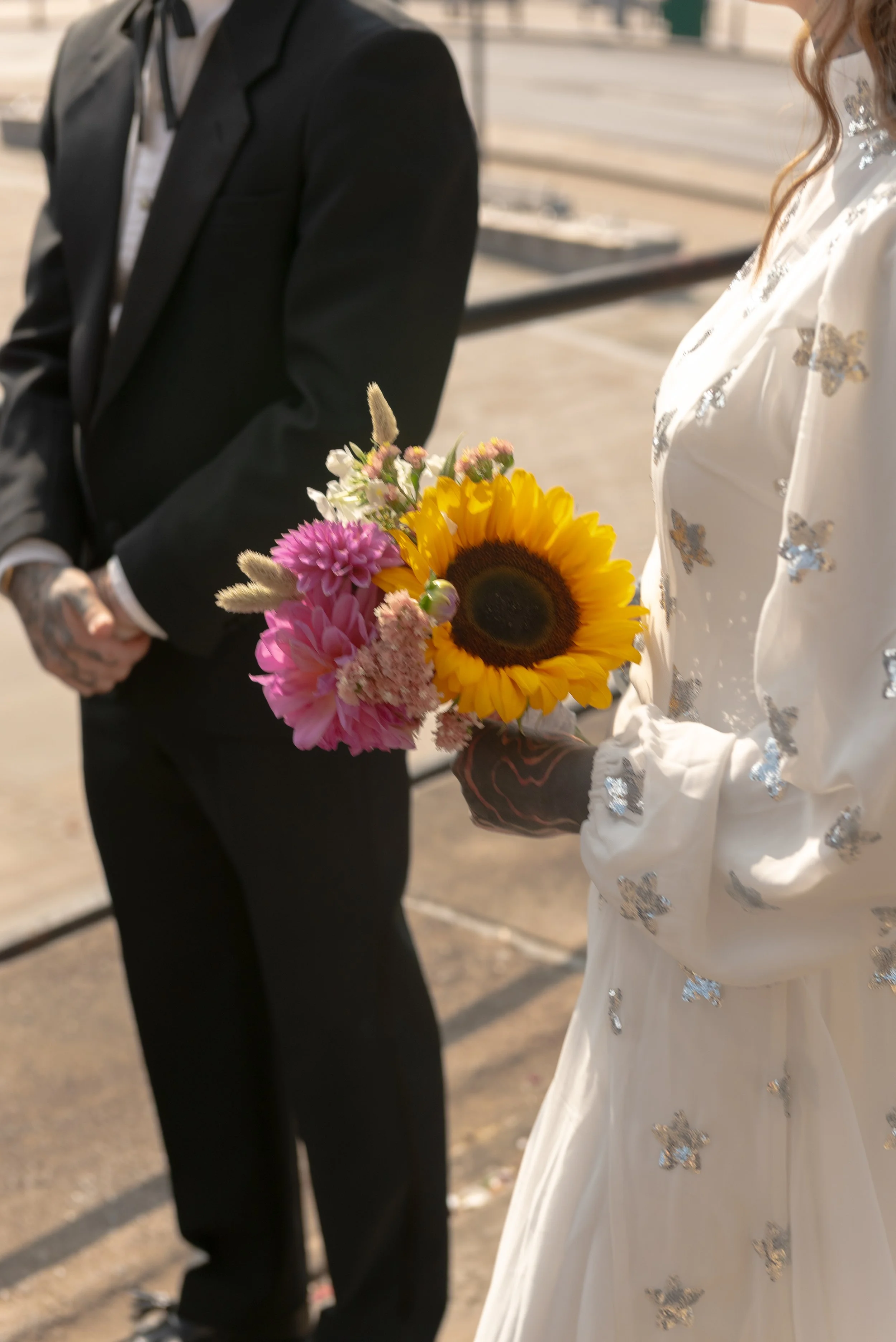 Person wearing a white dress with butterfly patterns holding a bouquet of sunflowers, pink dahlias, and other flowers, standing next to a person in a black suit with tattoos visible on their hands, outdoors on a sunny day.