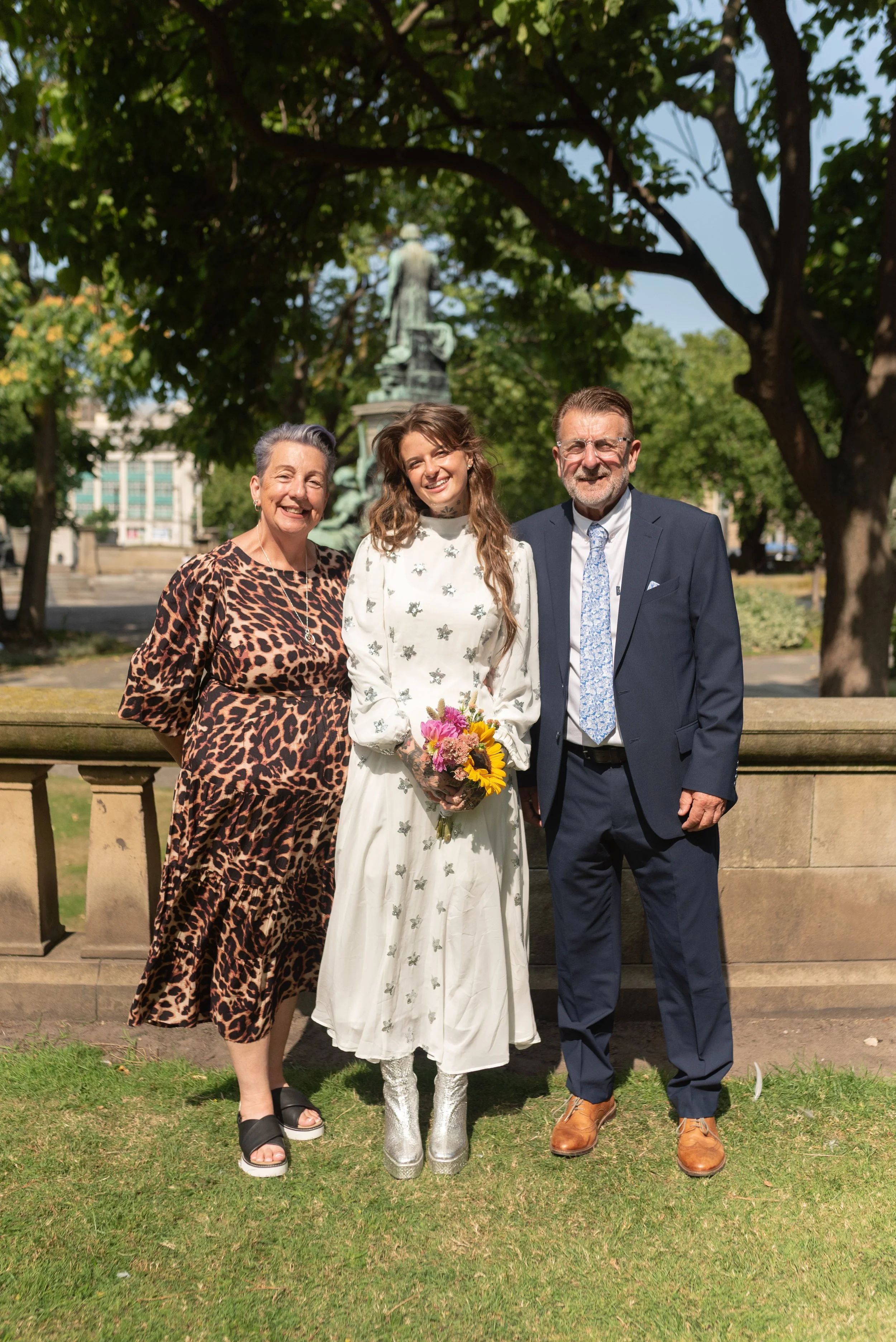 Group of three people standing outdoors in front of a stone railing, with a statue and trees visible in the background, smiling at the camera. The woman in the center is holding a bouquet of flowers.