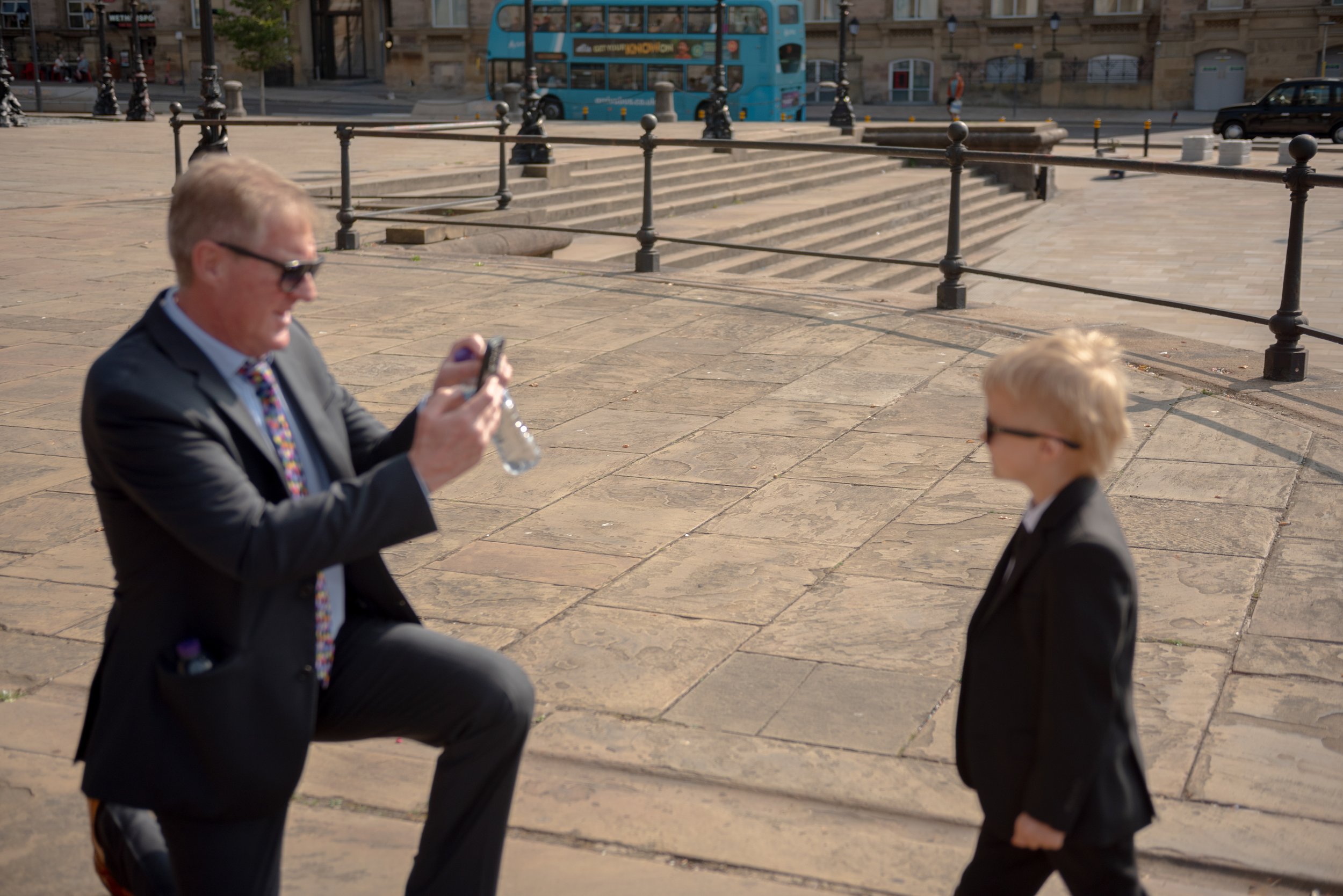 A man in a suit takes a photo of a young boy in a suit on a paved city sidewalk with steps and railings behind them, and street in the background.