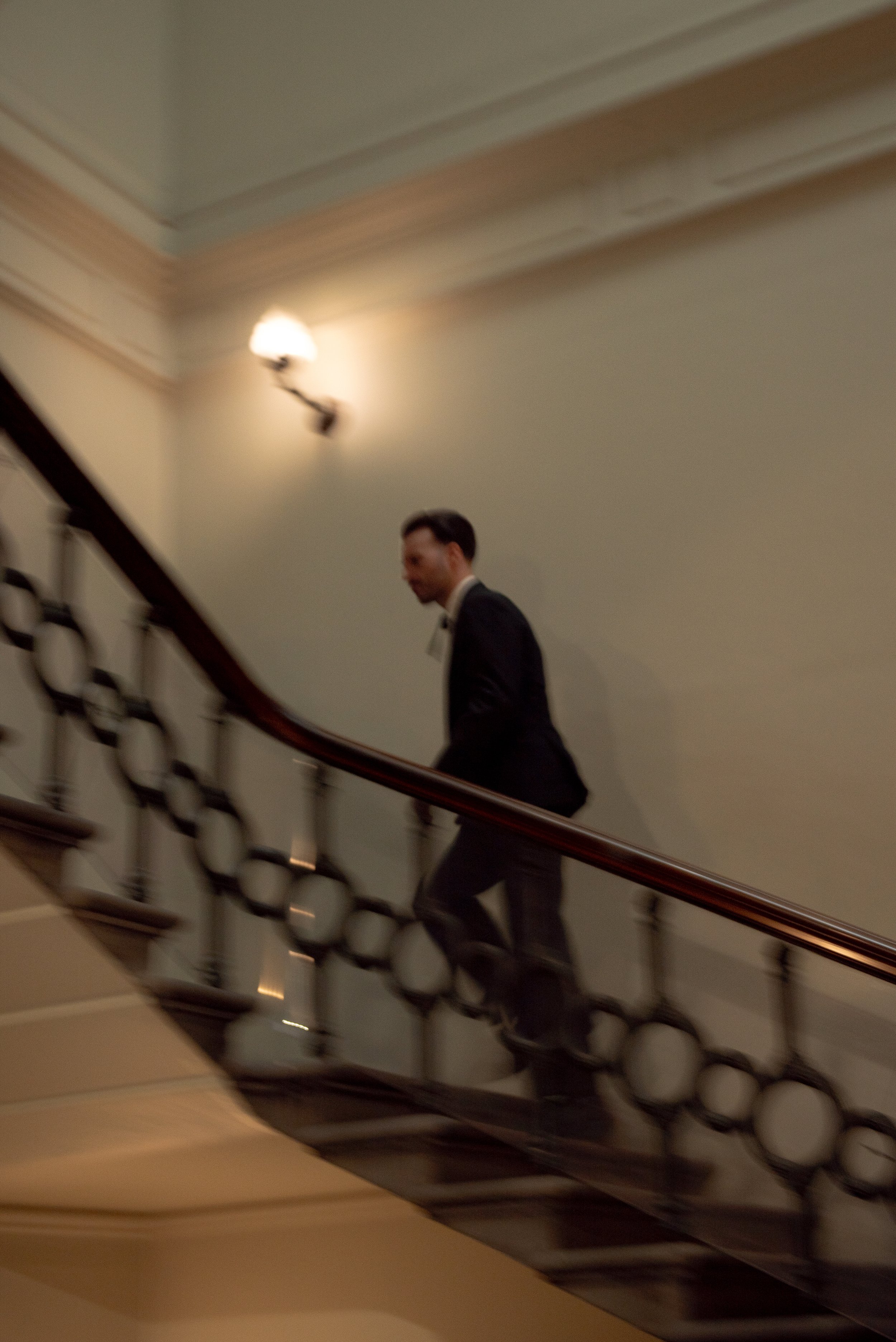A man in a tuxedo walking up a staircase with a decorative black and white railing, in a building with beige walls and a wall-mounted light fixture.