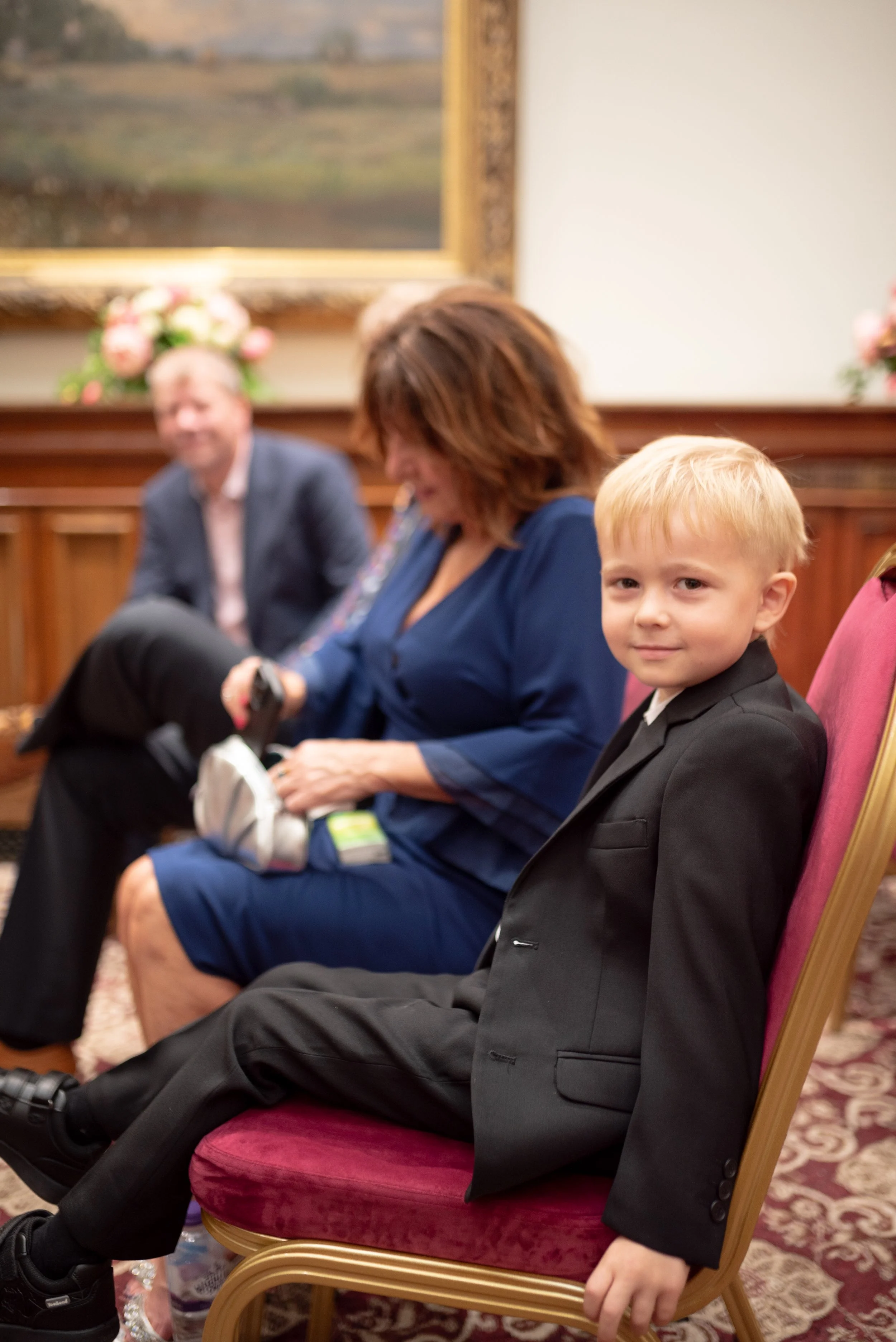 A young boy in a black suit sitting on a pink upholstered chair, looking at the camera, in a formal indoor setting with other adults seated nearby.