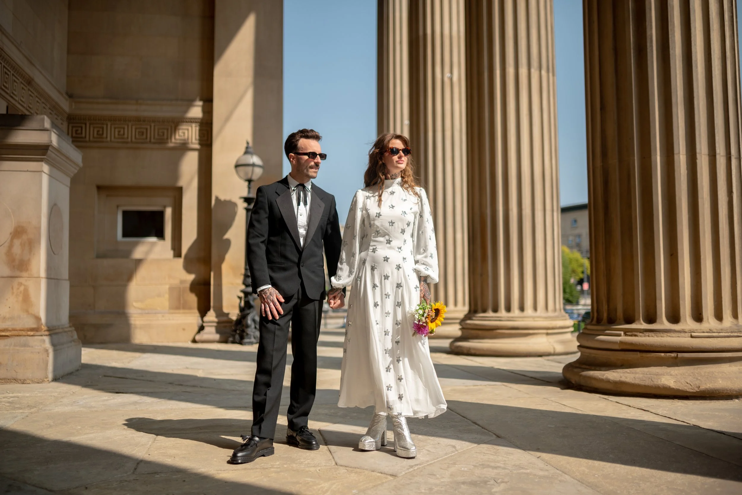 A couple in wedding attire standing hand in hand outside on a sunny day, surrounded by large classical columns of a historic building.