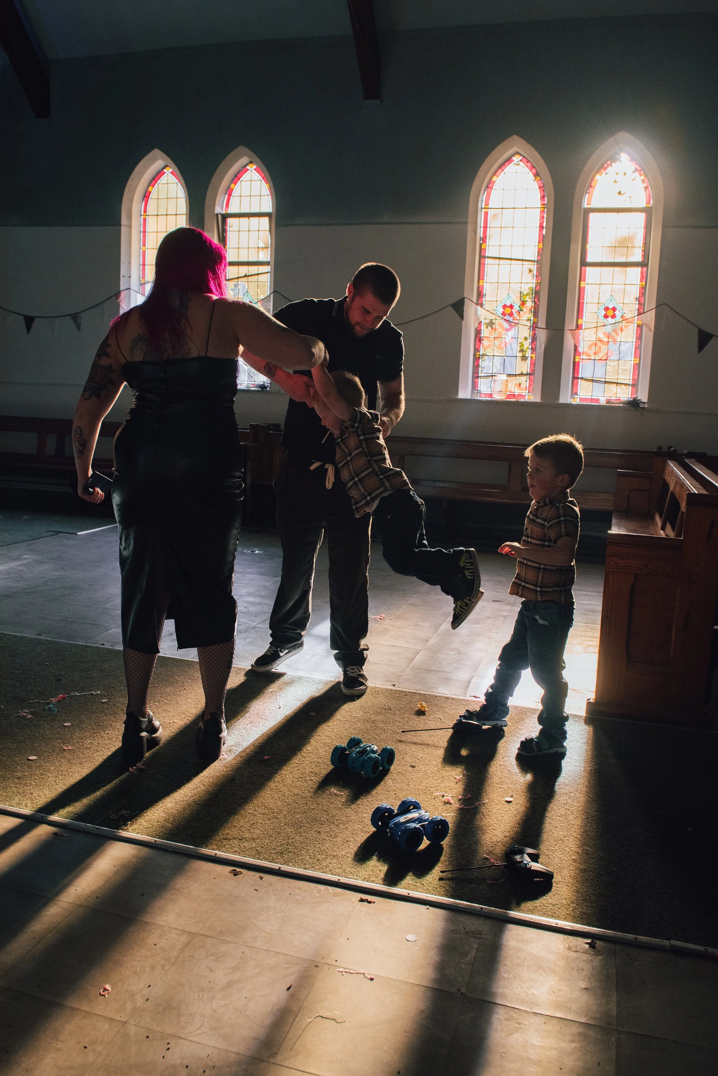 Inside a church with stained glass windows, a family is playing with remote-controlled cars on the floor, with sunlight streaming through the windows and casting shadows.