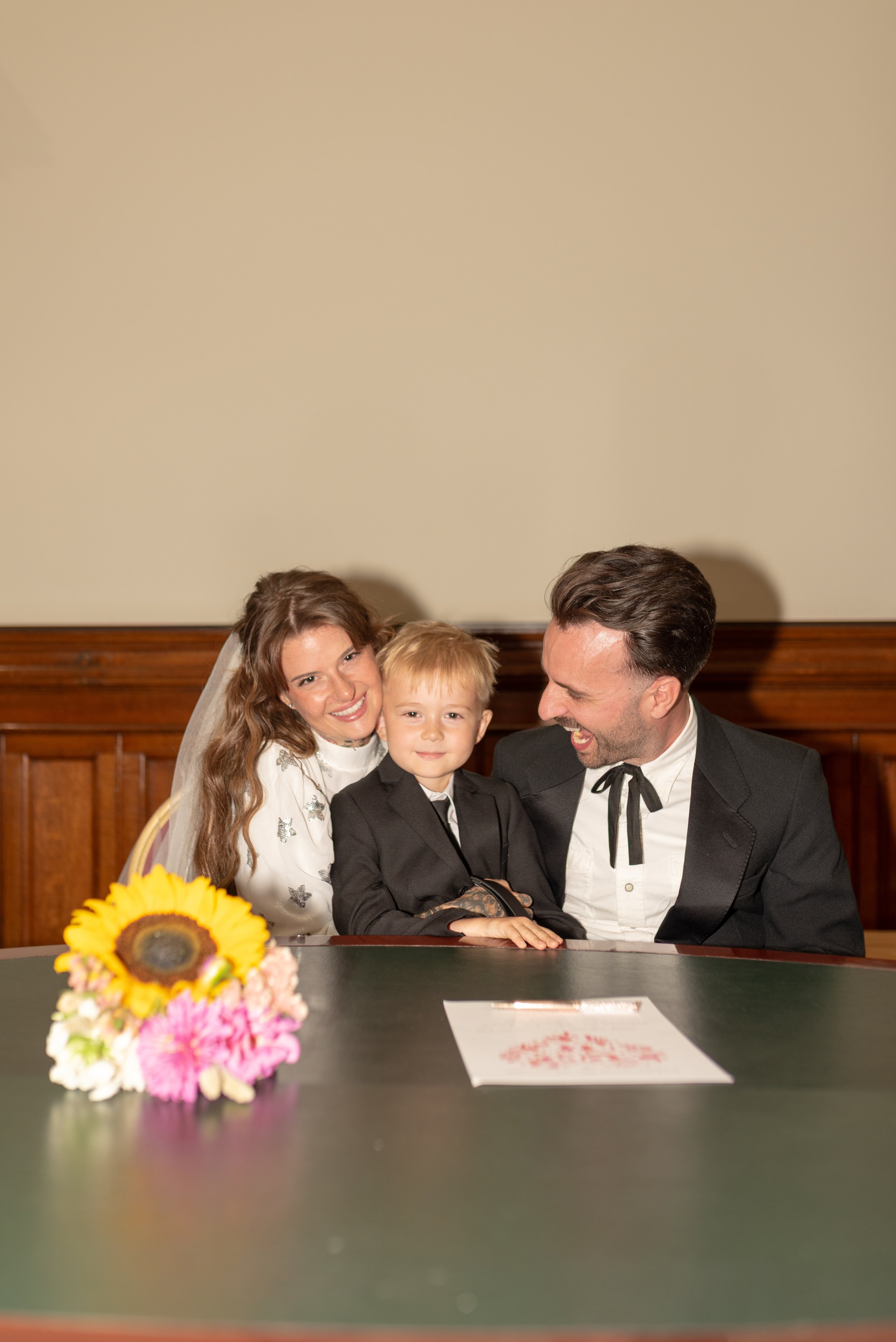 A family, including a woman, a young boy, and a man, are sitting together at a table in a room with wooden paneling. They are smiling and enjoying their time together, with a large sunflower and pink flowers on the table.