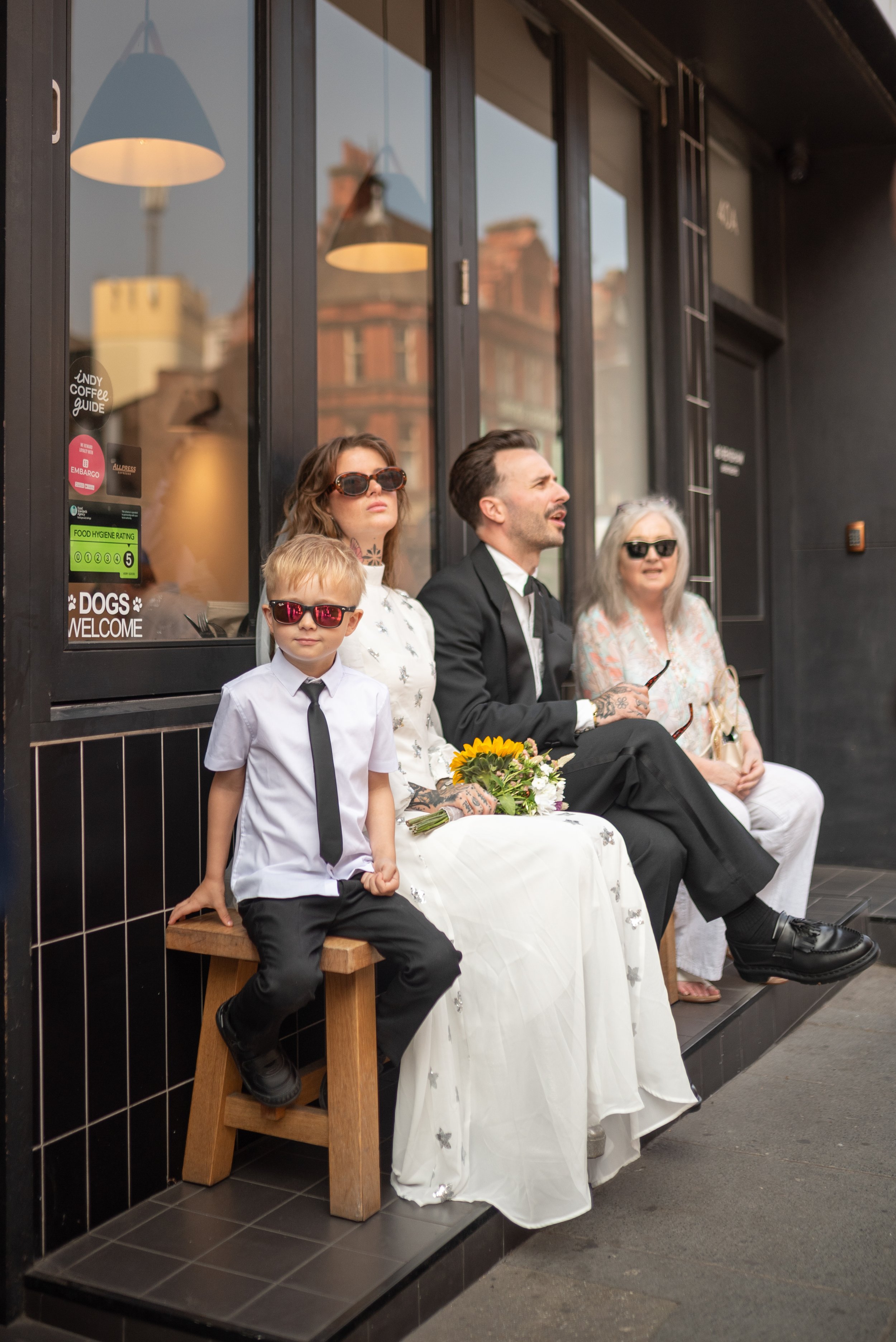 A group of four people sitting on a bench outside a cafe. They are dressed in formal and casual attire, with the woman in a white dress holding a bouquet of flowers, and all wearing sunglasses. The background shows the cafe window and a city street.