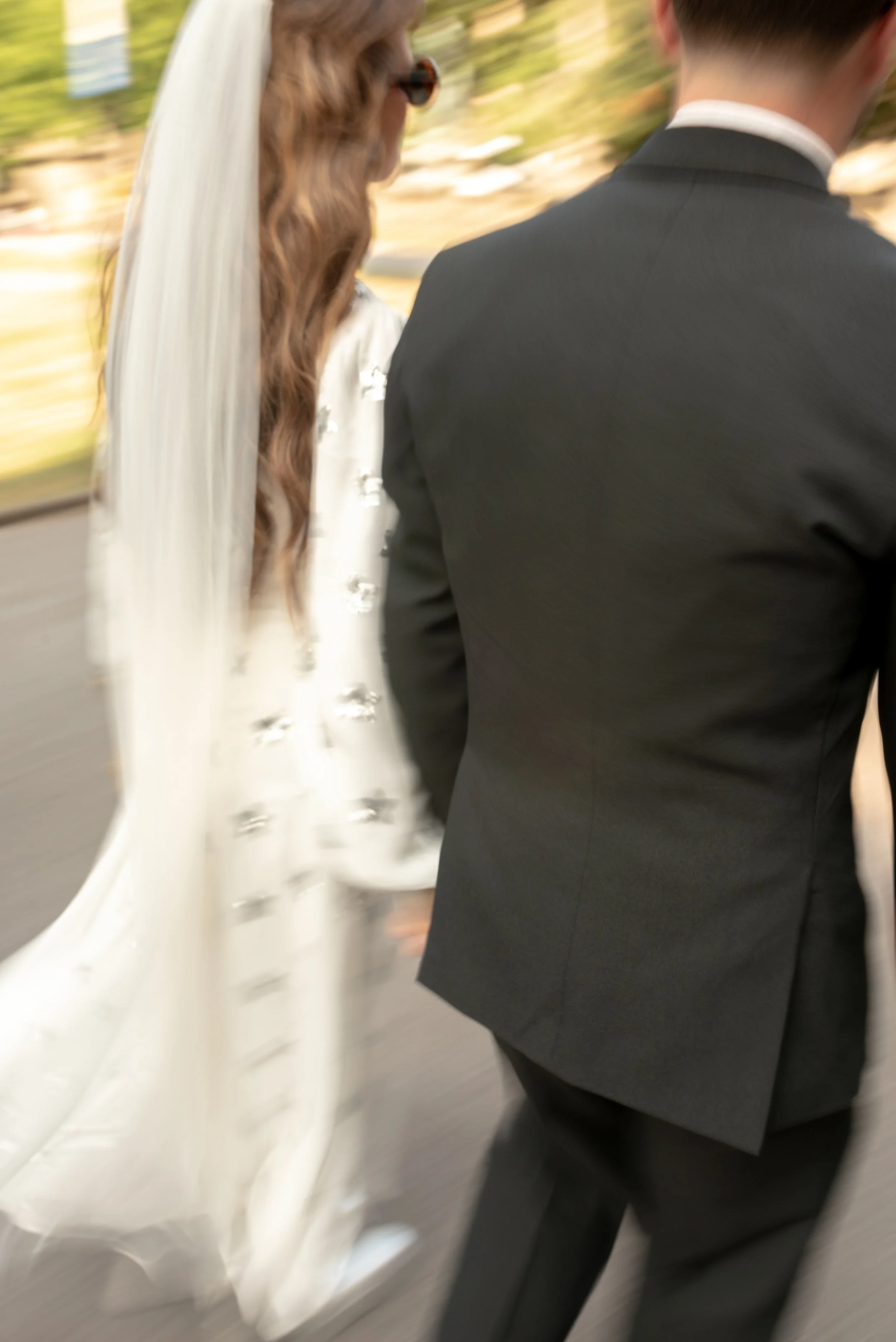 Blurred image of a bride and groom walking, seen from behind, with the bride in a white dress and veil, and the groom in a black tuxedo, outdoors.