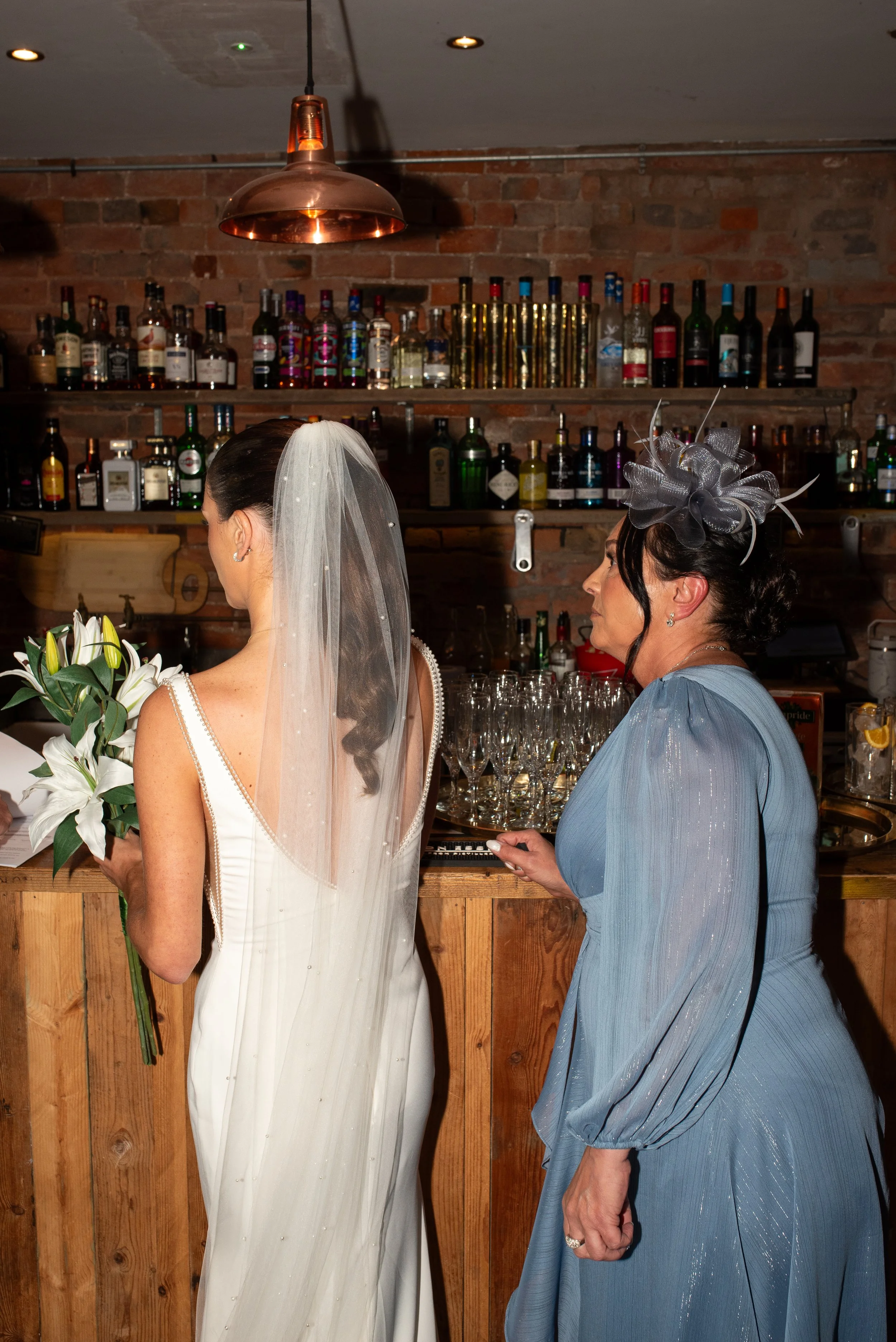 A bride holding a bouquet of white lilies, standing at a bar with a woman in a blue dress and hat nearby, against a brick wall background with bottles of alcohol.