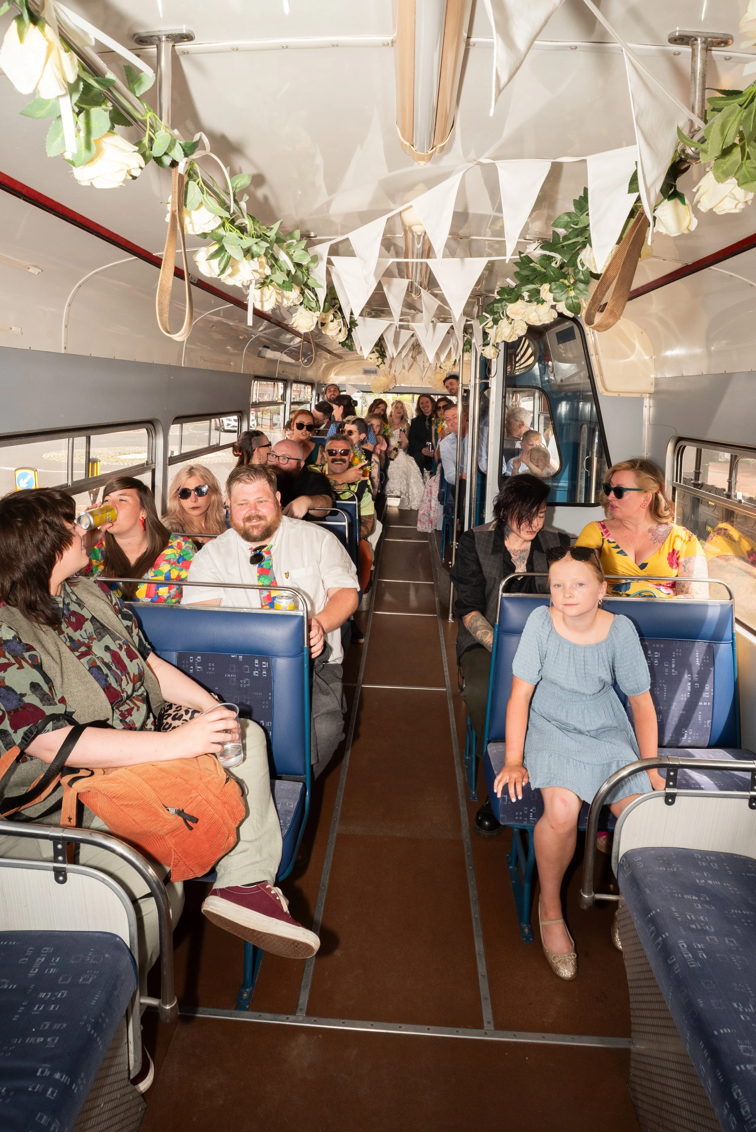 People celebrating on a decorated bus with white bunting and floral garlands, some wearing sunglasses, holding drinks, and smiling.