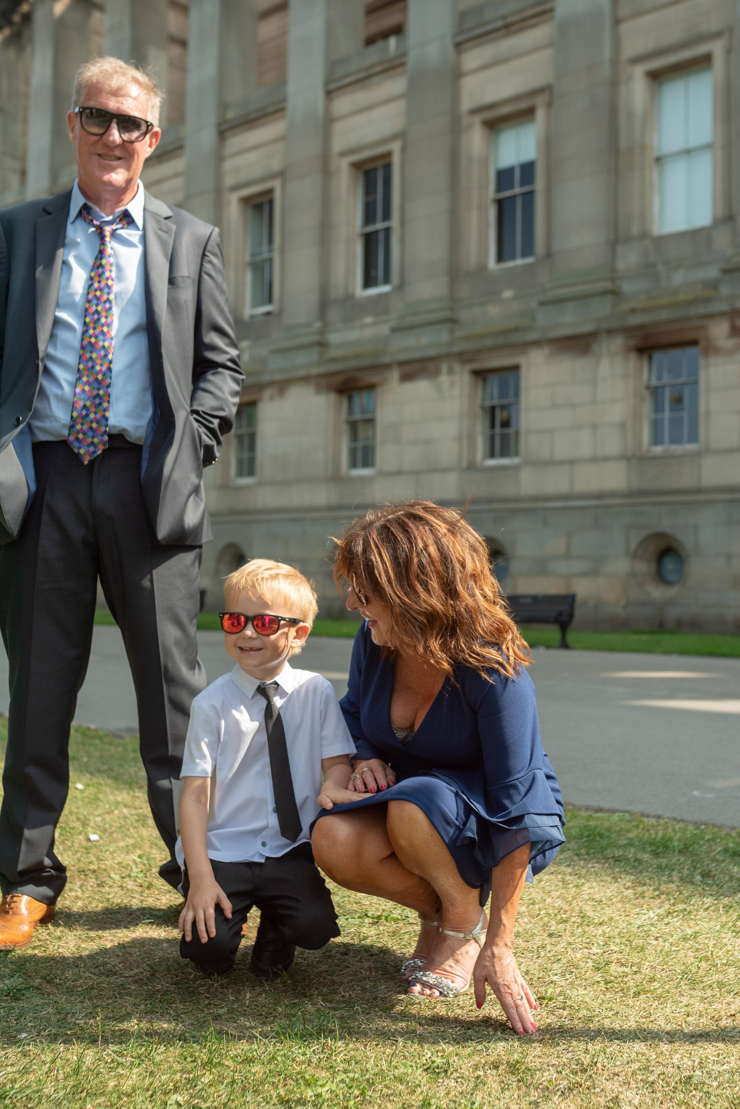 A woman crouching down on a grassy area next to a young boy wearing sunglasses, a white shirt, and a black tie, both smiling. An older man in a suit and colorful tie stands nearby, smiling and wearing sunglasses. They are outside in front of a large 