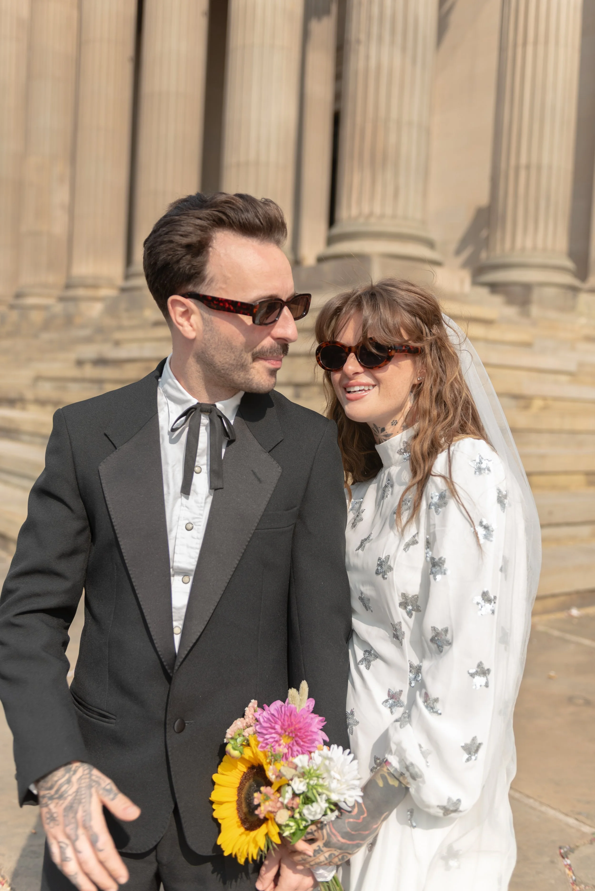 A couple dressed in wedding attire standing on stairs in front of a classical building with tall columns. The woman is holding a bouquet with sunflowers and pink flowers, wearing sunglasses and a white dress with silver butterfly embellishments. The 