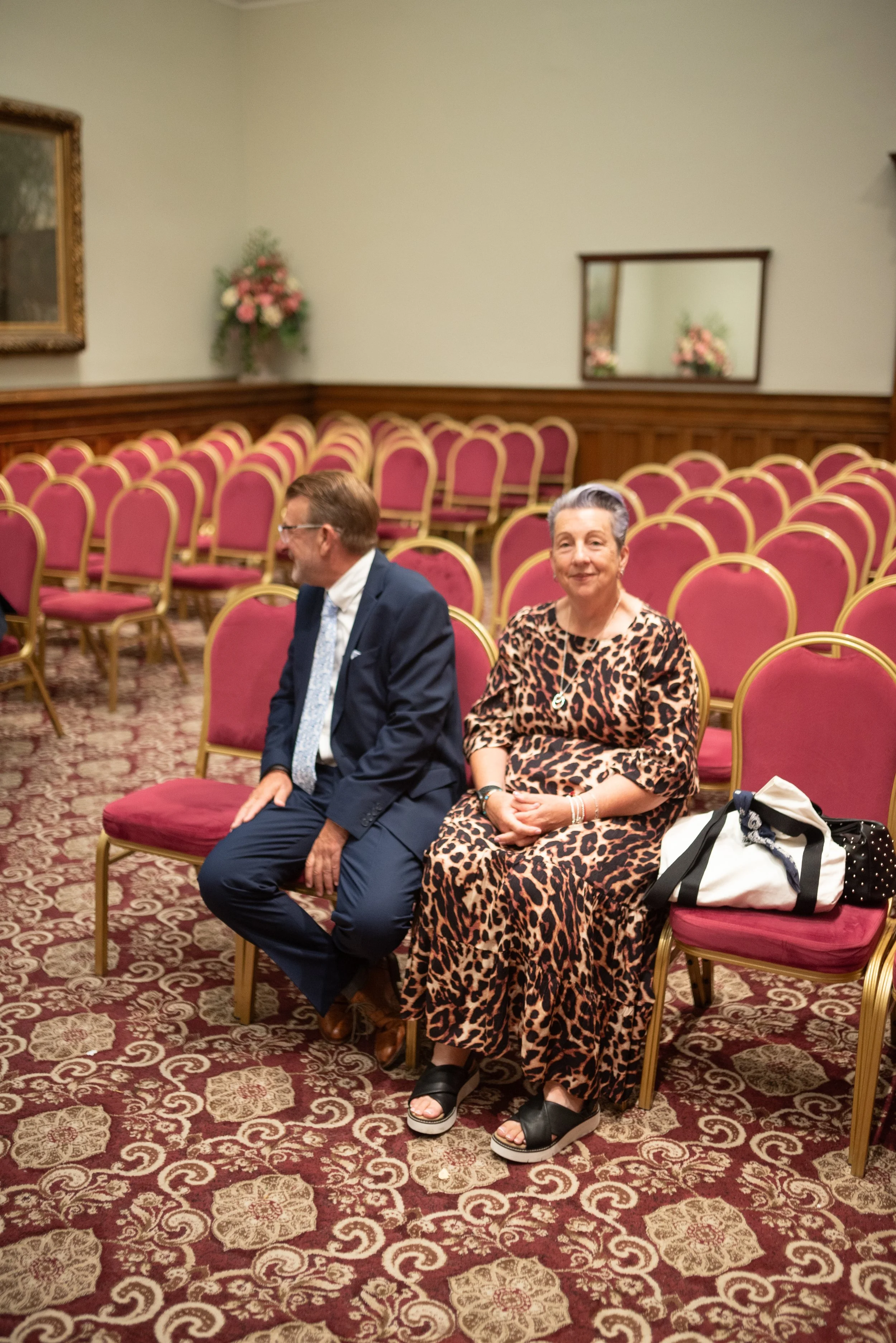 A man in a navy suit and a woman in a leopard print dress sitting on empty chairs in a decorated room with pink and gold chairs, floral arrangements, and a mirror on the wall.