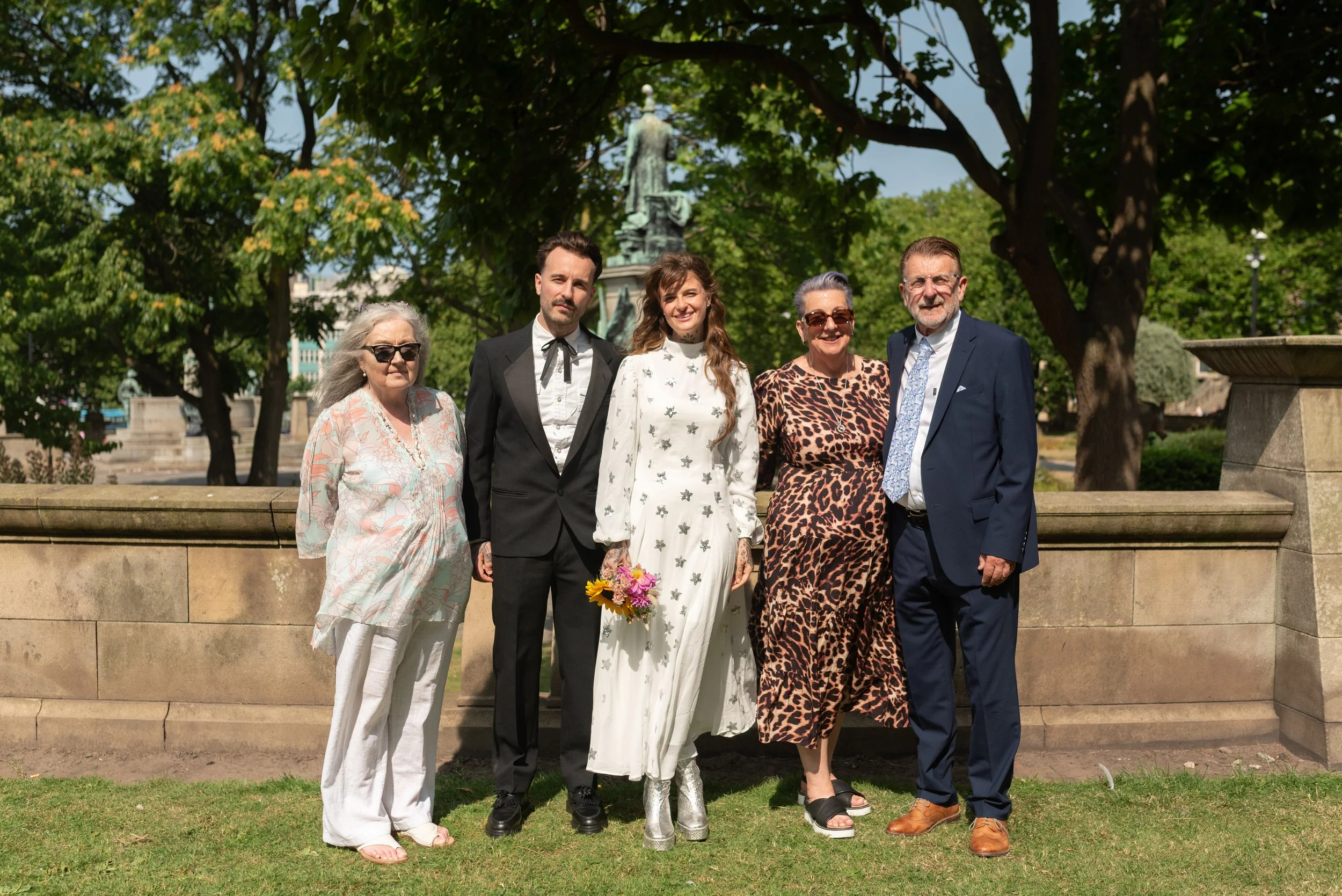Group of six people standing outdoors on a sunny day in front of a stone wall with trees and a monument in the background, dressed in formal and casual attire, possibly for a celebration or event.