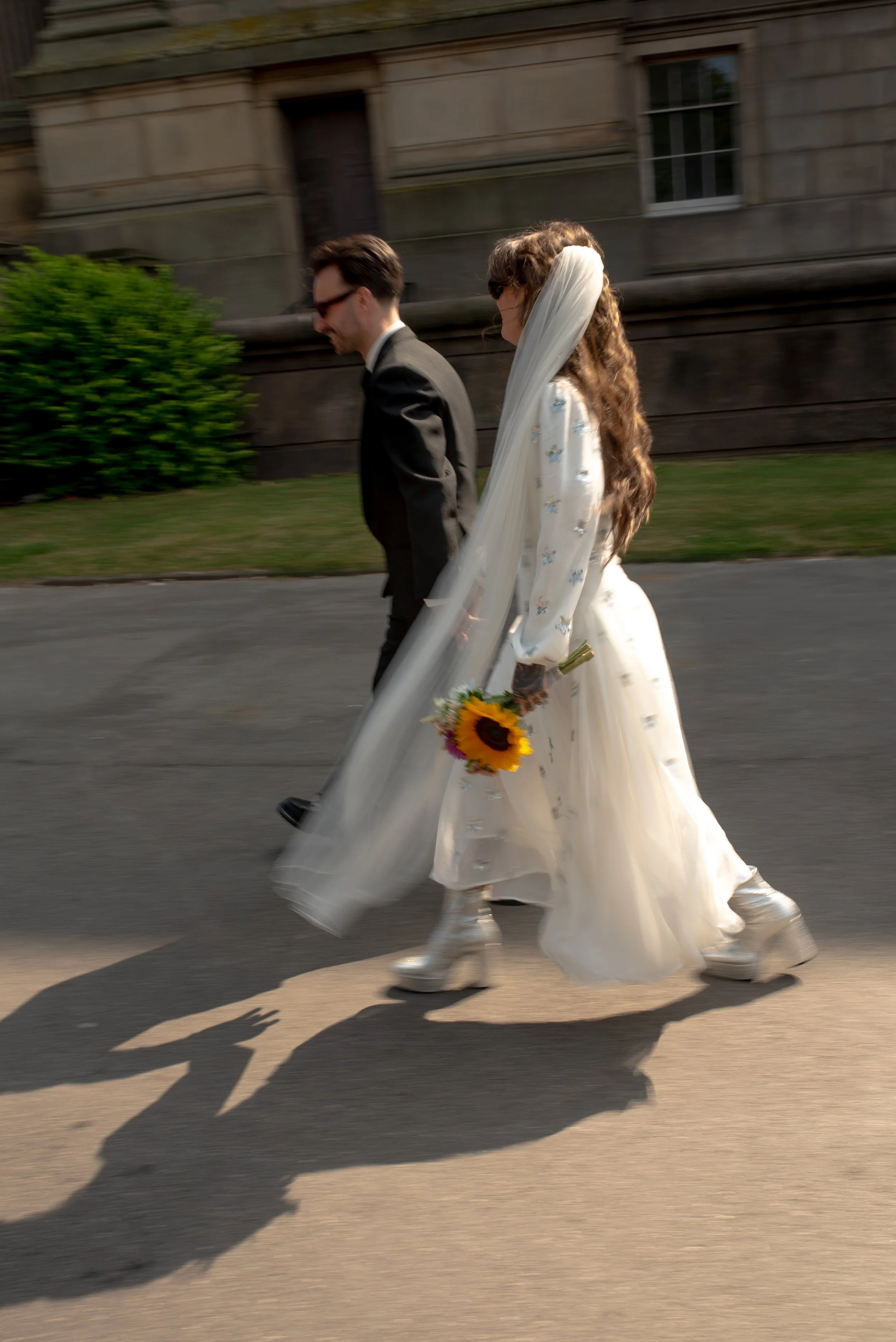 A bride and groom walk together on a street. The bride is holding a sunflower and wearing a dress with a long veil and platform boots. The groom is dressed in a suit and glasses. Their shadows are cast on the pavement.