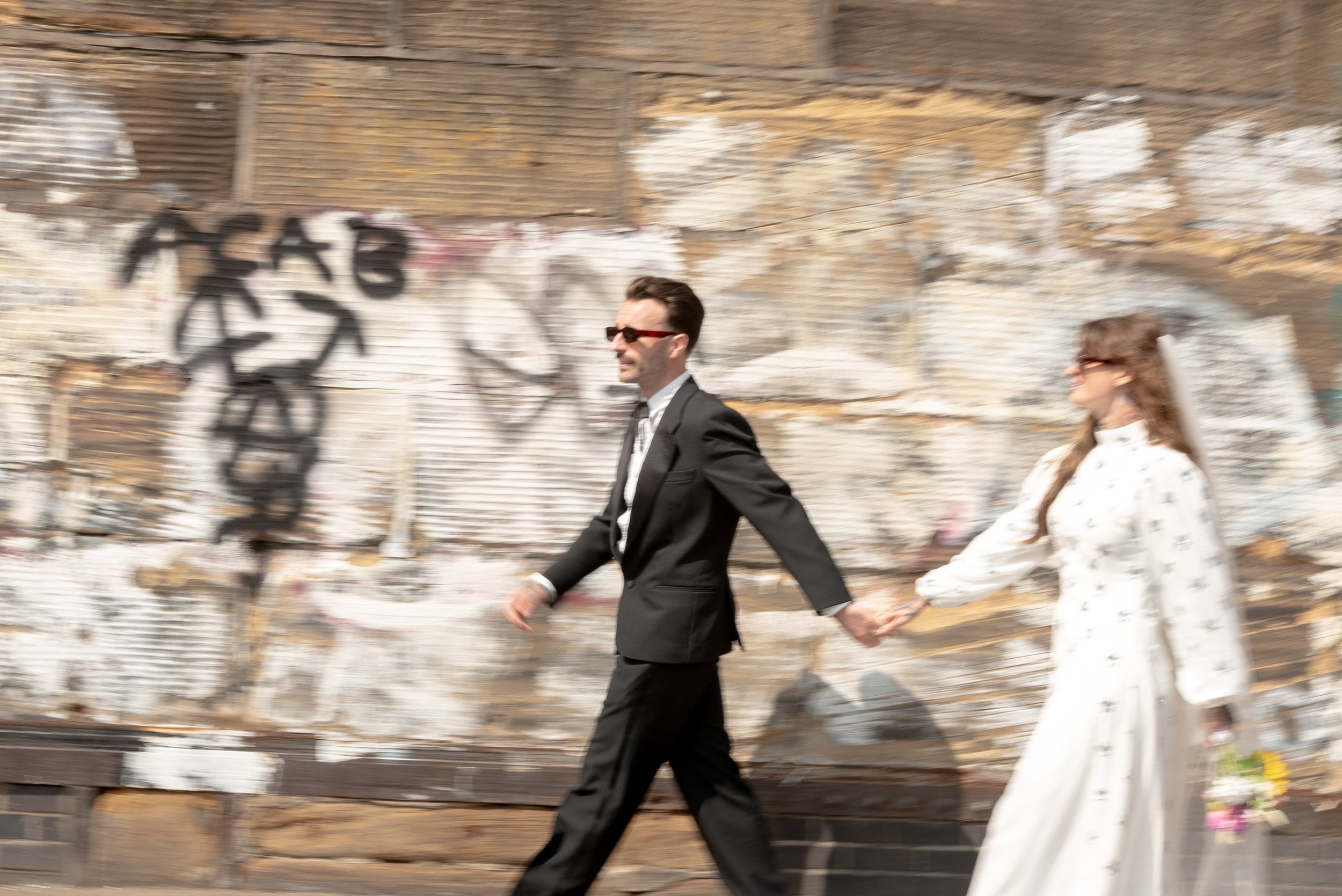 A man and woman holding hands while walking downtown, with a graffiti-covered wall in the background.