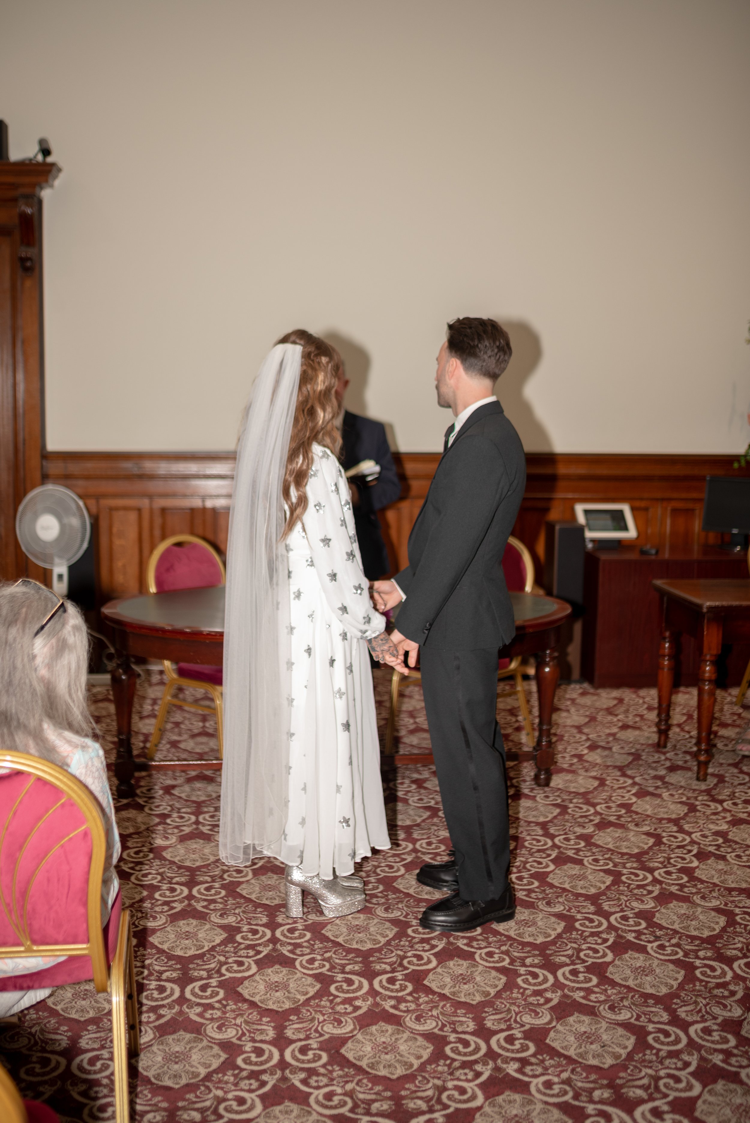 A bride and groom holding hands during their wedding ceremony, facing each other indoors with guests seated nearby.
