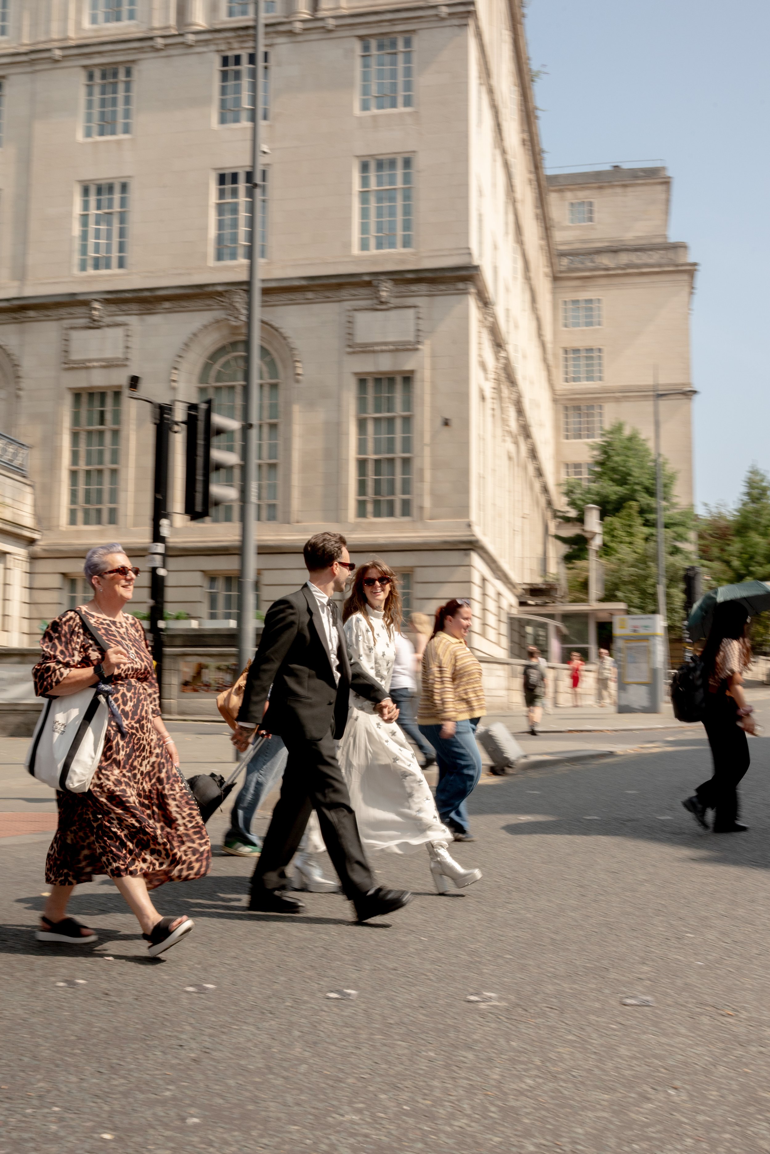 People crossing the street in front of a large, historic building on a sunny day.