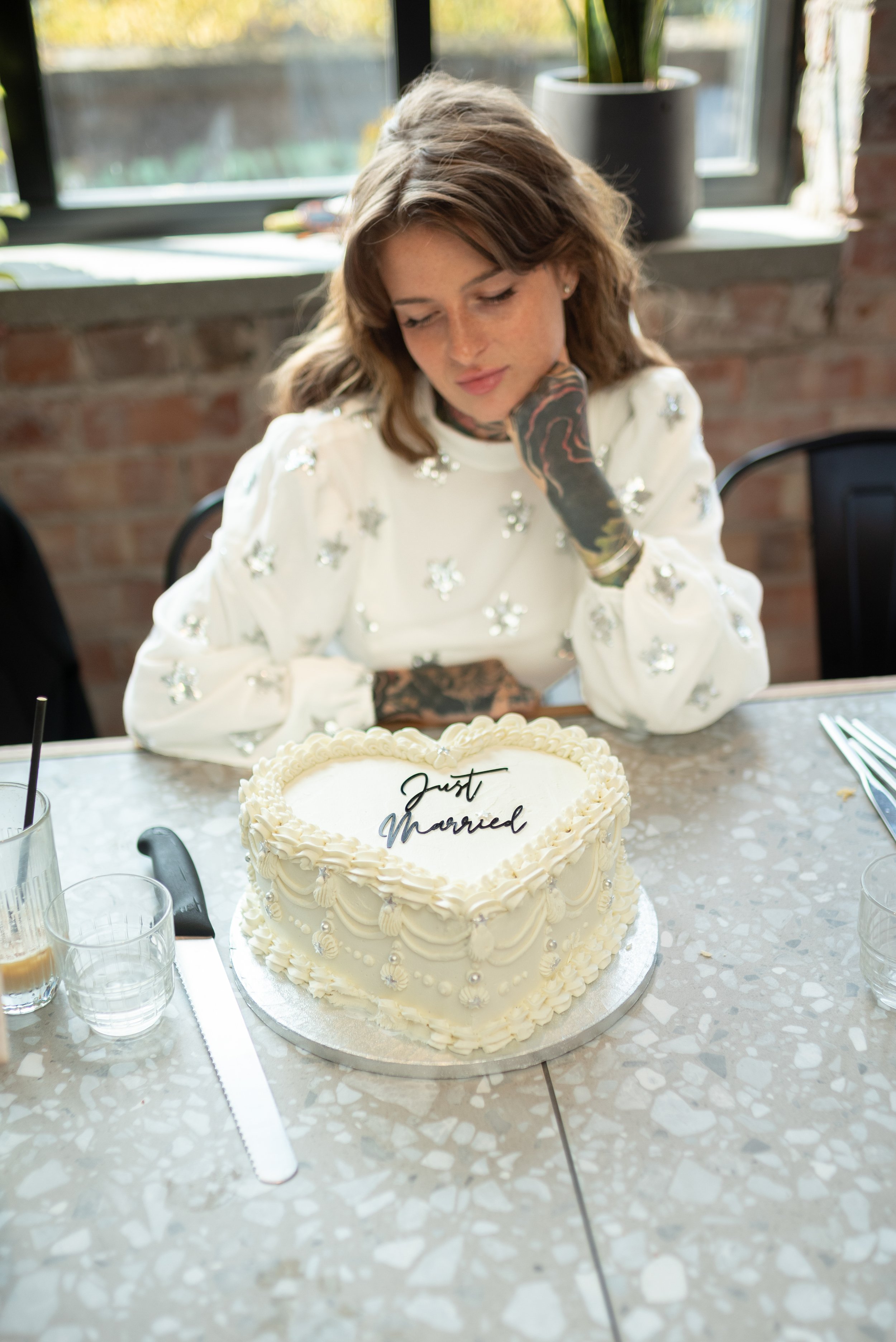 A woman with brown hair and tattoos, sitting at a table with her chin resting on her hand, looking at a heart-shaped wedding cake with "Just Married" written on top.