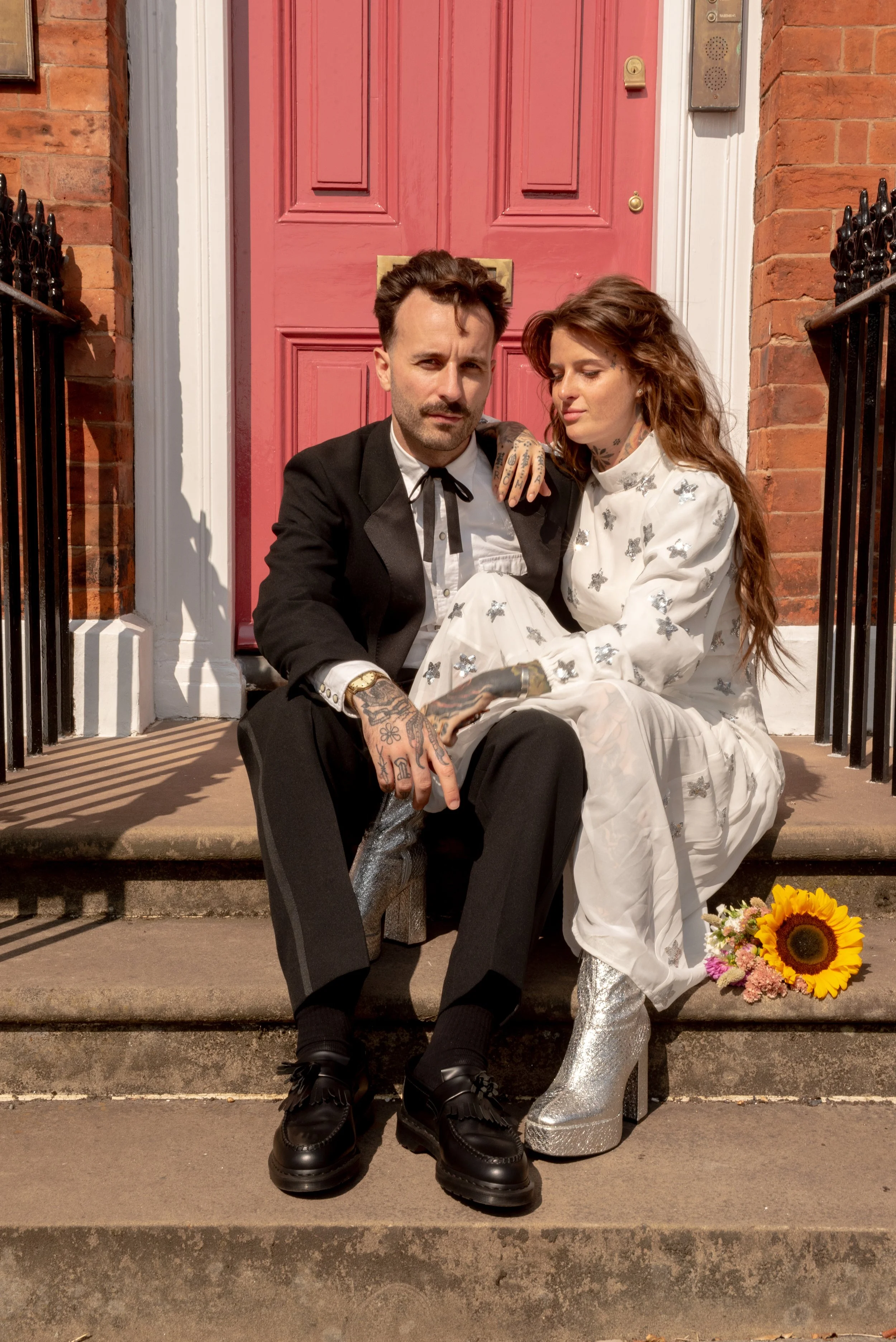 A man and a woman sitting on the steps of a porch in front of a red door. The man is in a black suit with tattoos and holding a silver boot. The woman is in a white dress with silver boots, holding a bouquet of flowers, with long red hair.