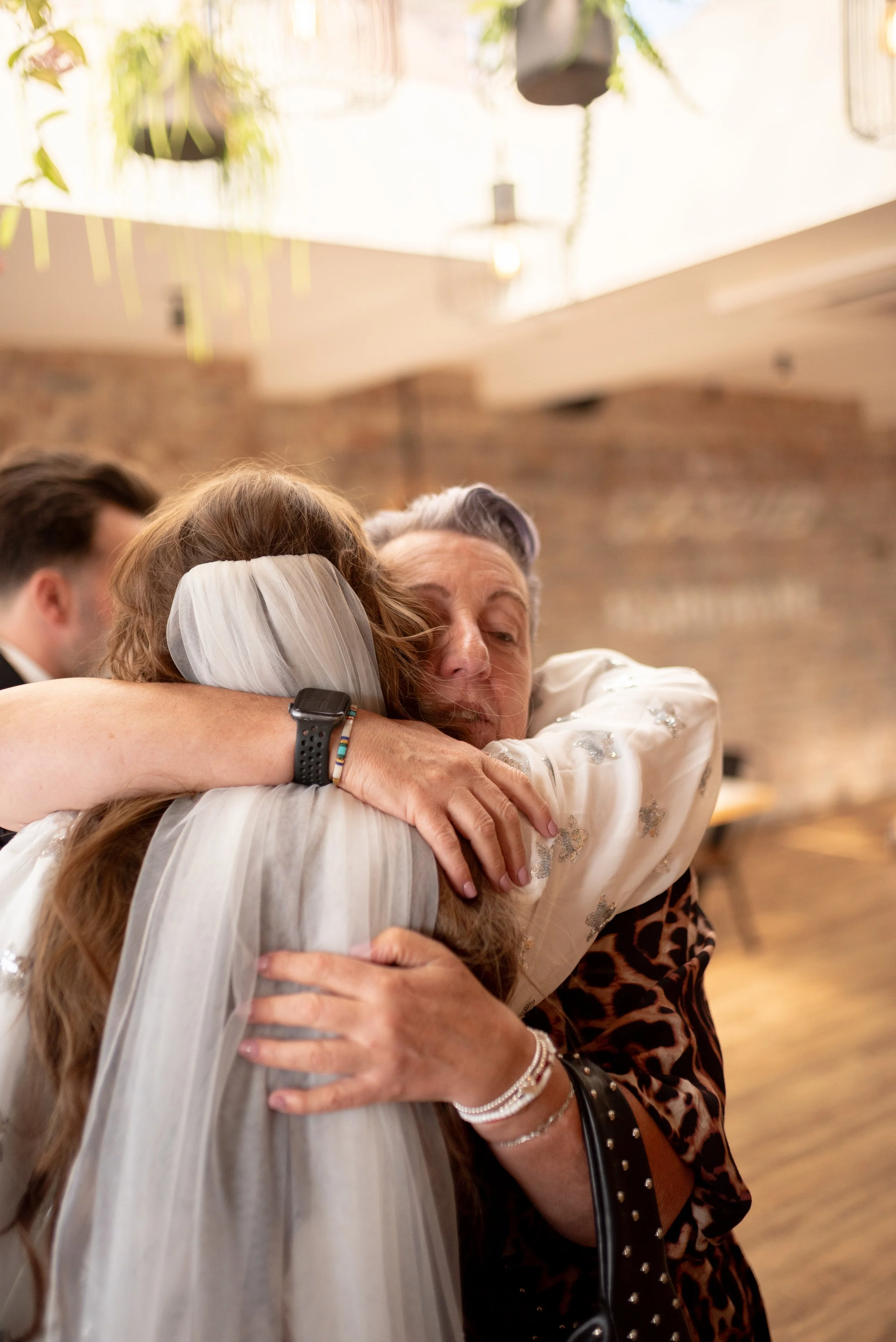 Two women hugging warmly at a celebration, one wearing a veil and the other with gray hair, in a warmly lit indoor setting.
