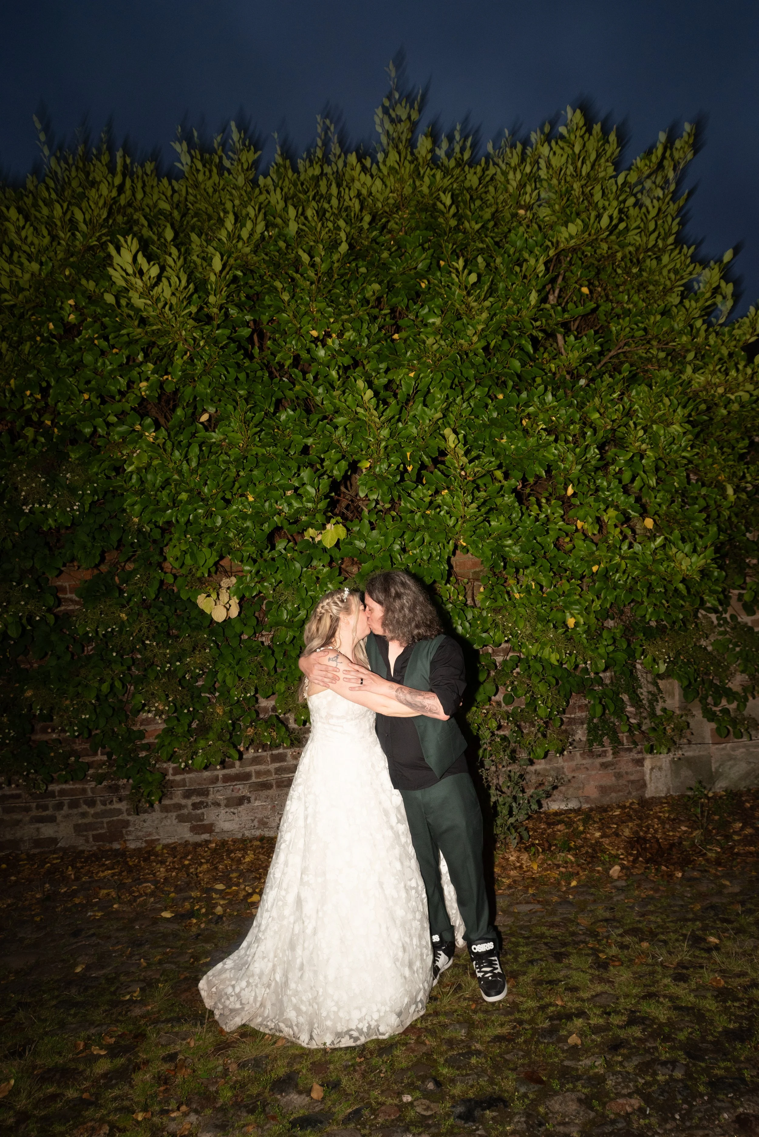 A couple kissing, the woman in a white wedding dress and the man in dark clothing, standing in front of a large green bush at night.