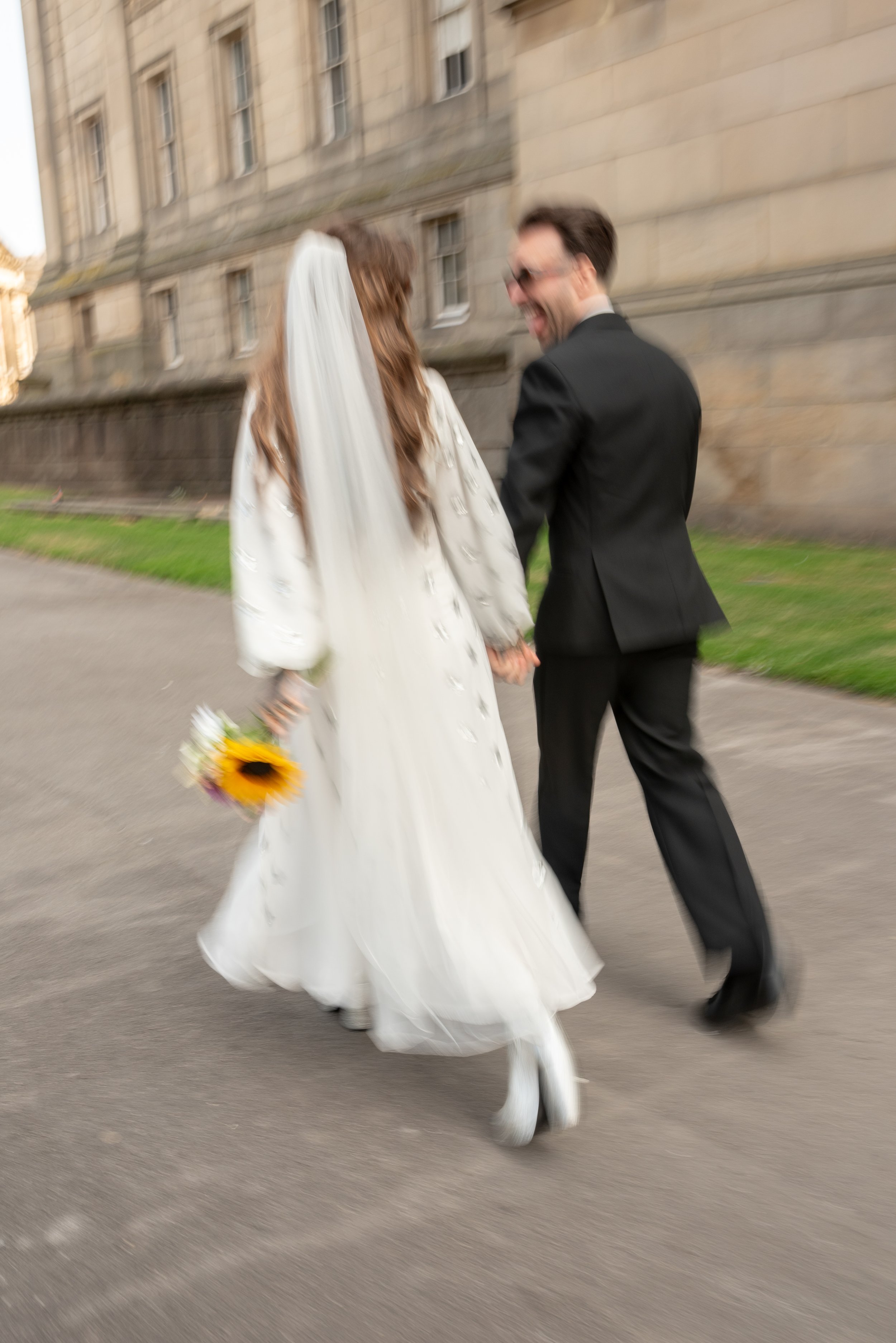 Blurred photo of a bride and groom holding hands and walking outdoors on a sidewalk, with the bride holding a sunflower bouquet and wearing a white wedding dress and veil, and the groom in a black tuxedo, in front of a large stone building.