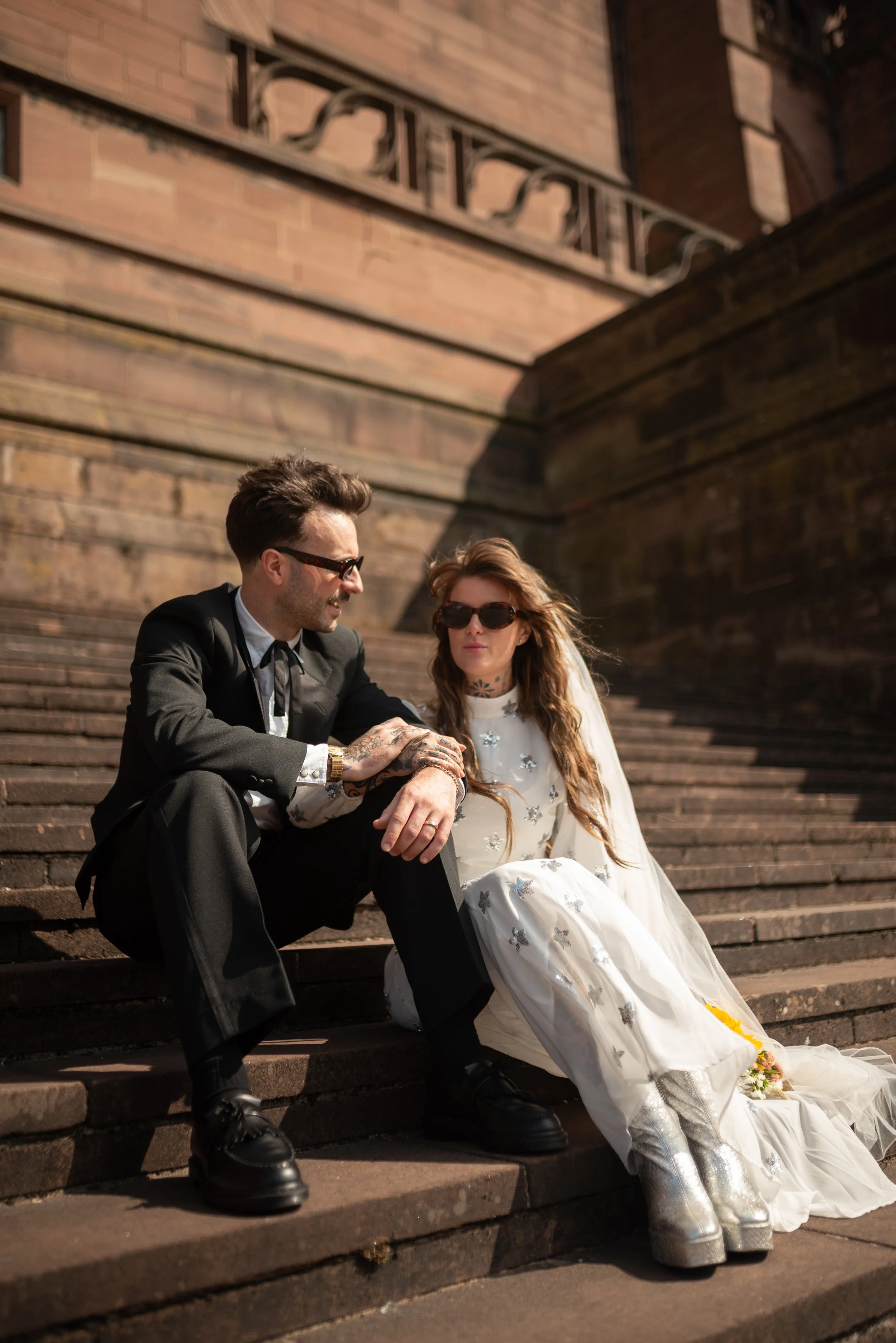 A man and woman sitting on outdoor stone stairs, dressed in wedding attire, with the man in a black suit and the woman in a white dress with silver boots, both wearing sunglasses, in a sunny setting.