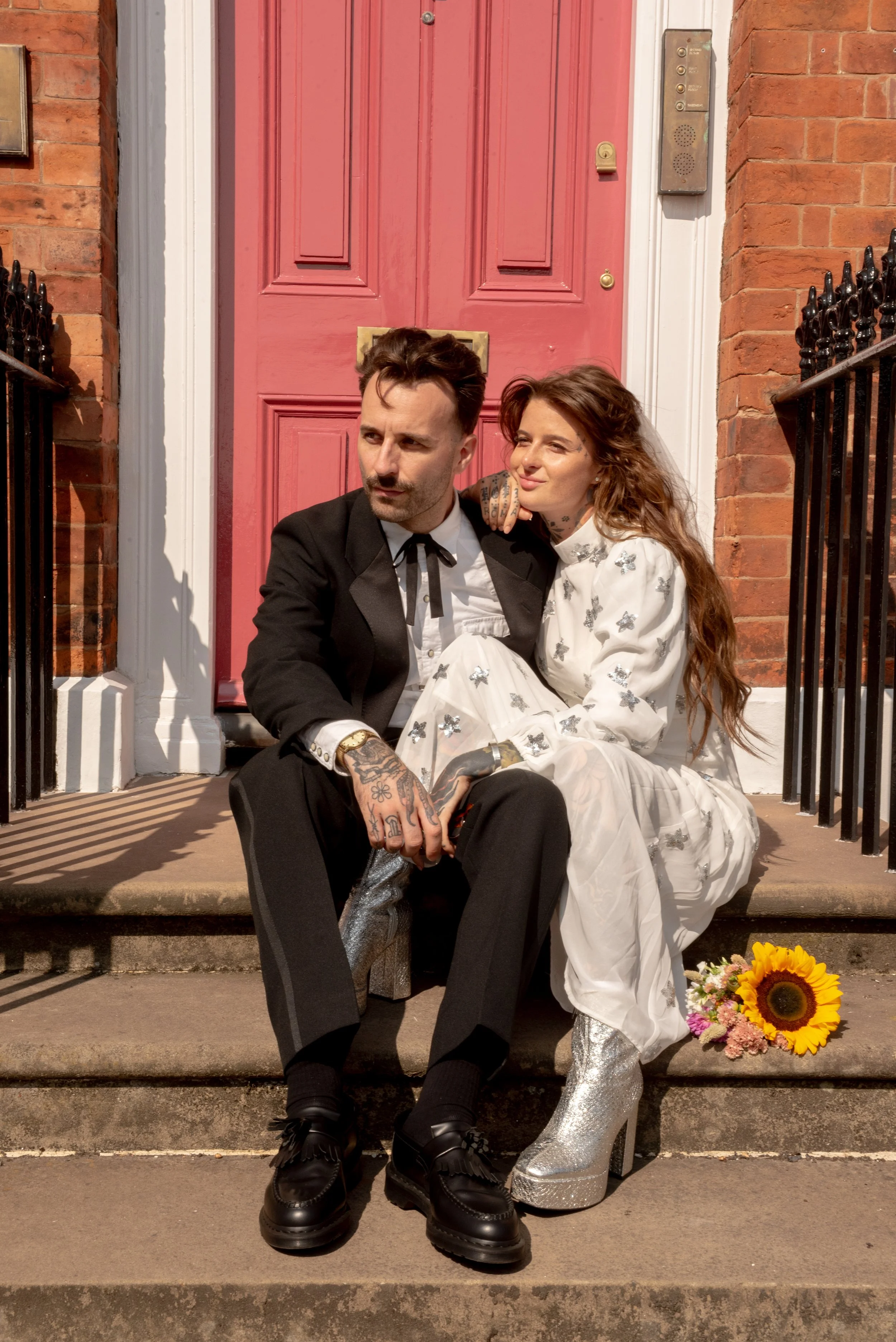 A man and woman sitting on front steps of a red door, holding hands. The man is dressed in a black suit, and the woman is wearing a white dress with a butterfly pattern and sparkly silver high-heeled boots. A sunflower and pink flowers are on the ste