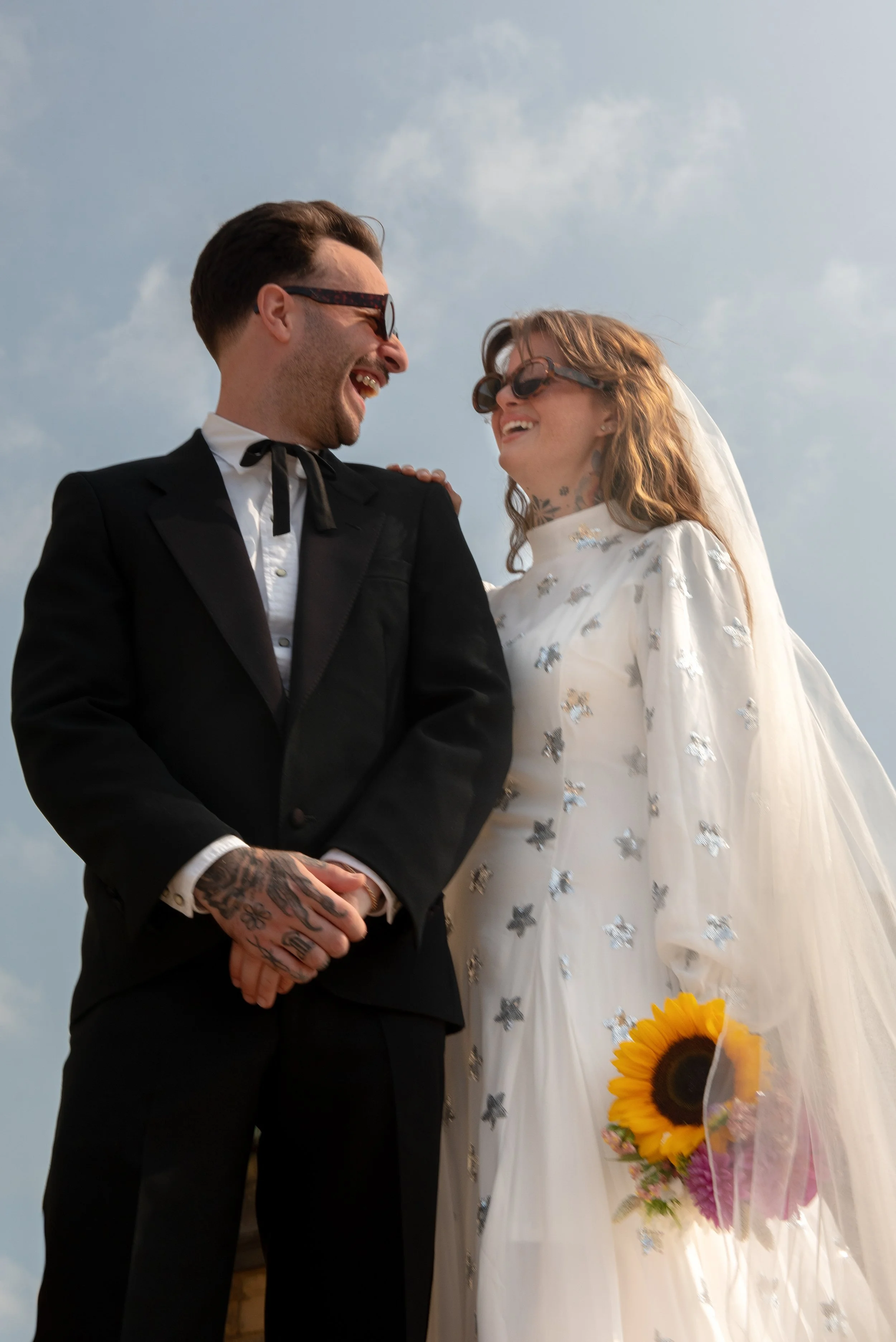 Happy couple on their wedding day, holding hands and smiling at each other. The groom is in a black suit with tattoos on his hands, and the bride is wearing a white dress with silver star details, holding a sunflower bouquet, both wearing sunglasses,