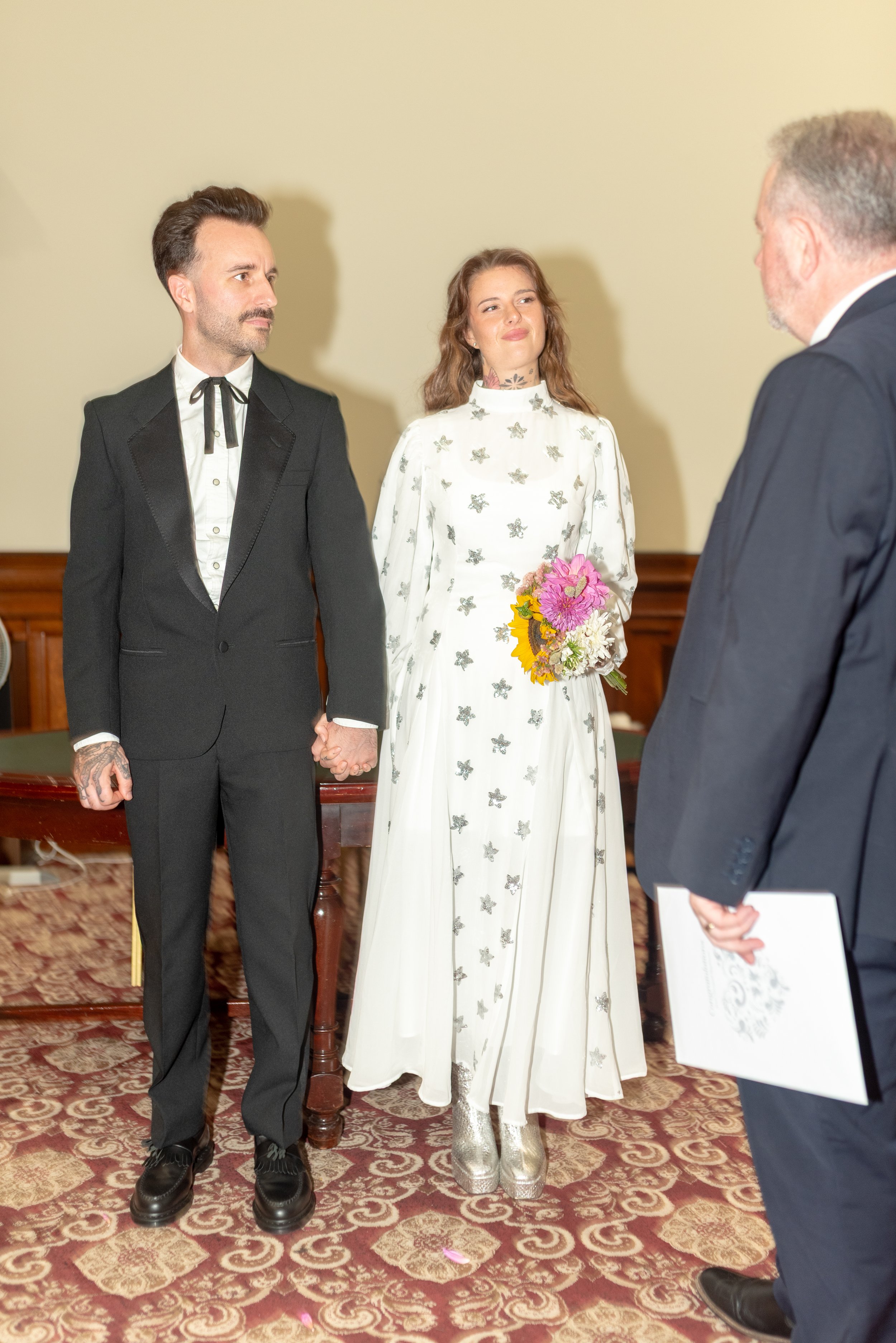 A couple holding hands during their wedding ceremony, with a woman in a white dress with silver butterflies and silver shoes, and a man in a black suit with a bow tie, facing an officiant in a dark suit. The woman is holding a bouquet of pink, yellow