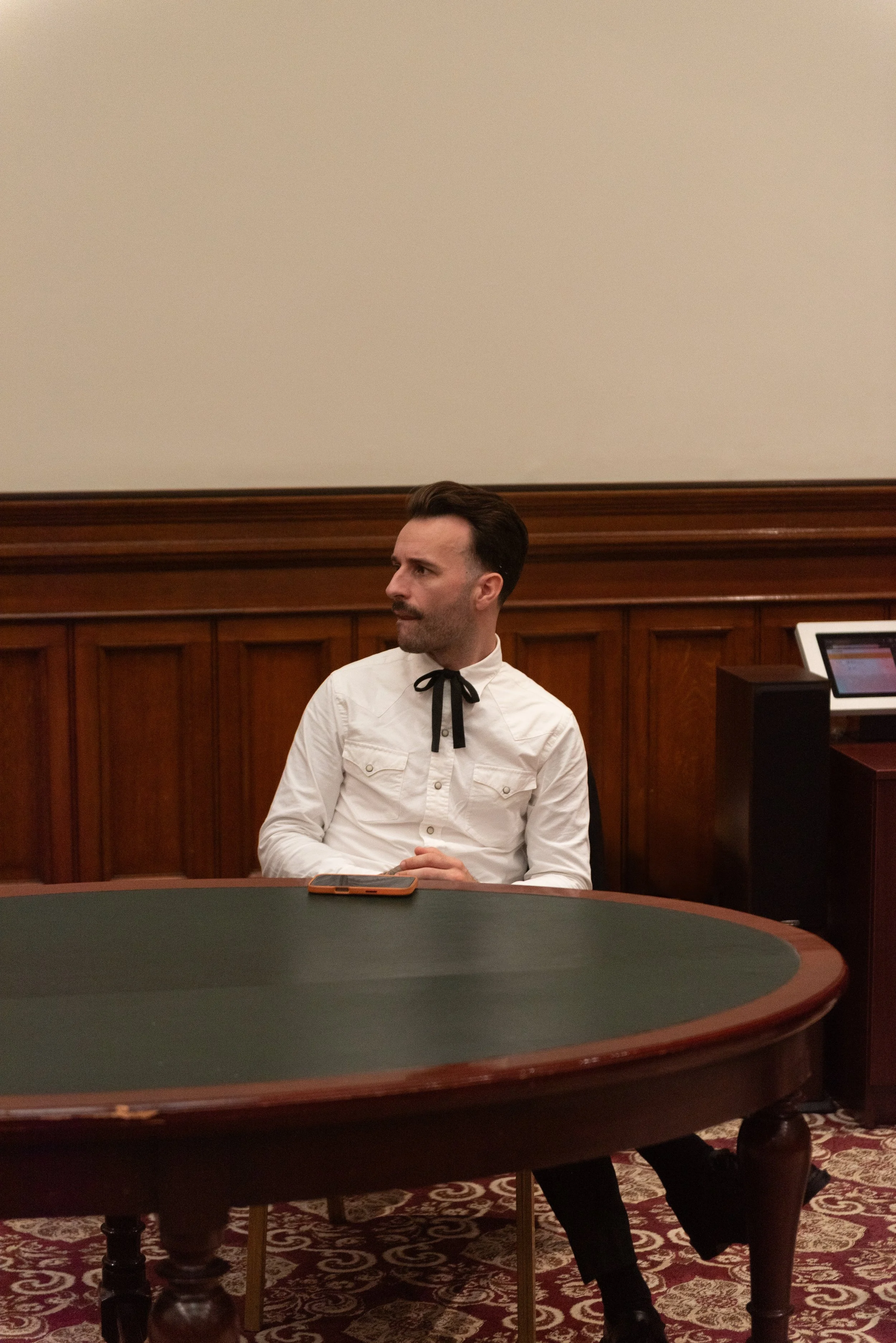 Man with dark hair and a beard, wearing a white button-up shirt with a black ribbon tie, sitting alone at a round card table in a room with wood-paneled walls, with a smartphone on the table.