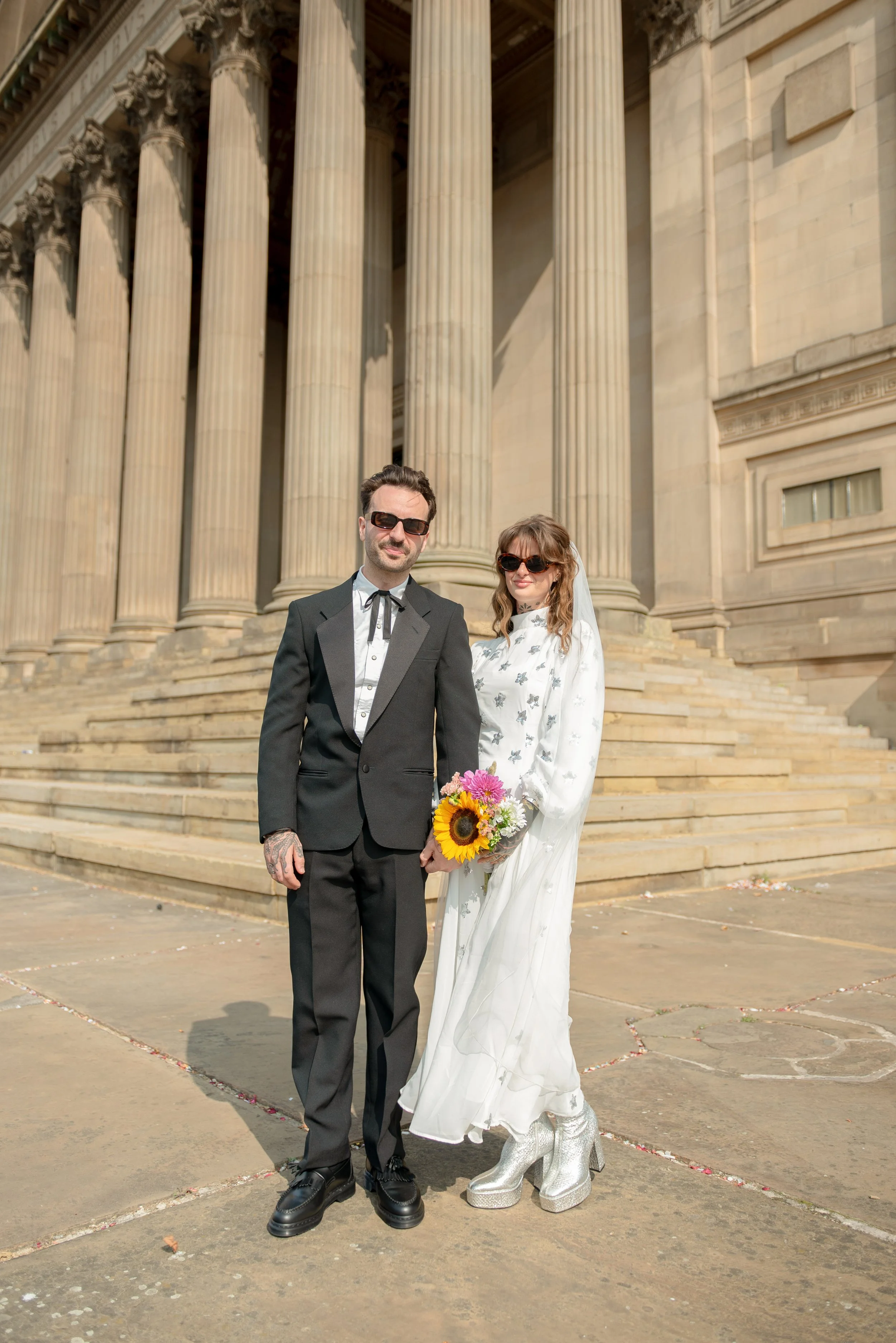 A couple dressed in wedding attire standing on the steps of a neoclassical building with large columns; the woman is holding a bouquet of flowers and wearing silver platform boots, and the man is wearing a black tuxedo with sunglasses.