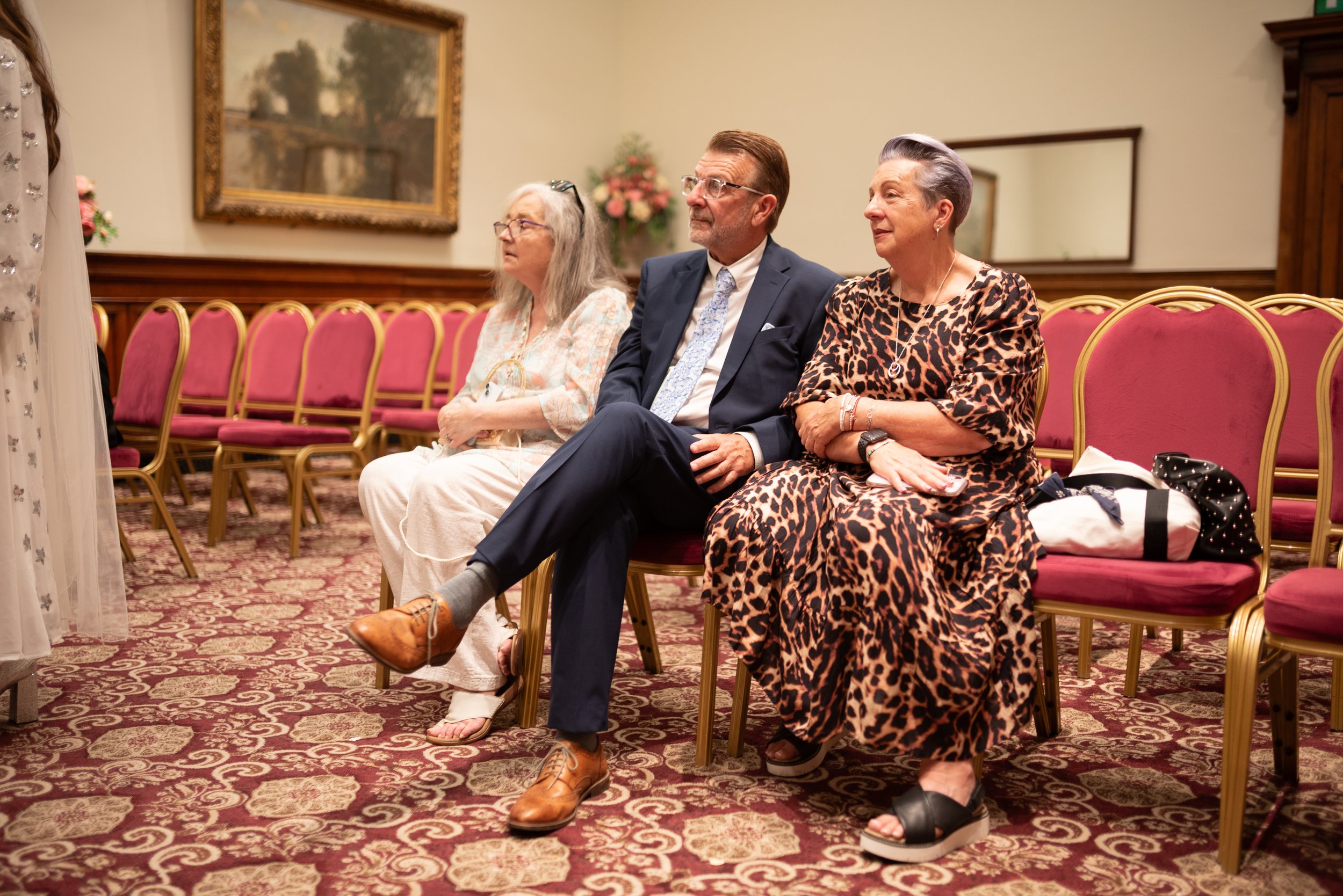 Three adults sitting in pink and gold chairs at an event, in a room with floral carpet, wood-paneled walls, and artwork, watching intently.