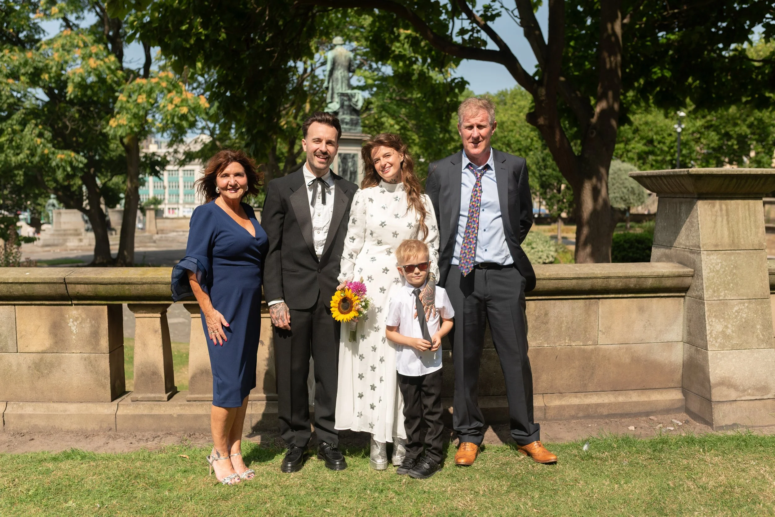 Group of six people dressed in formal and casual clothes standing outside in a park, smiling for a photograph, with trees and a statue in the background.