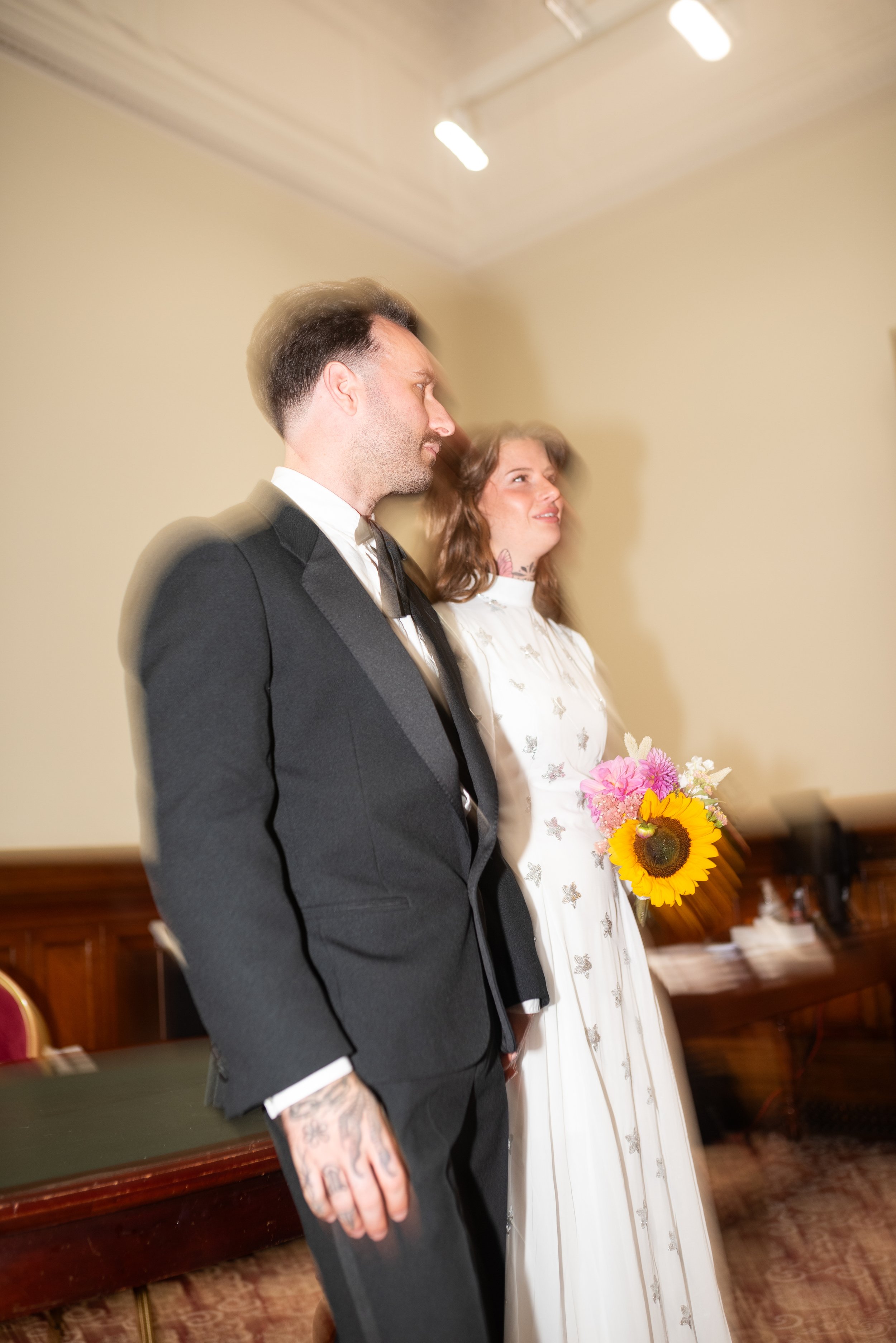 A bride and groom standing side by side in an indoor setting, with the bride holding a bouquet of flowers including a sunflower, and both looking ahead.