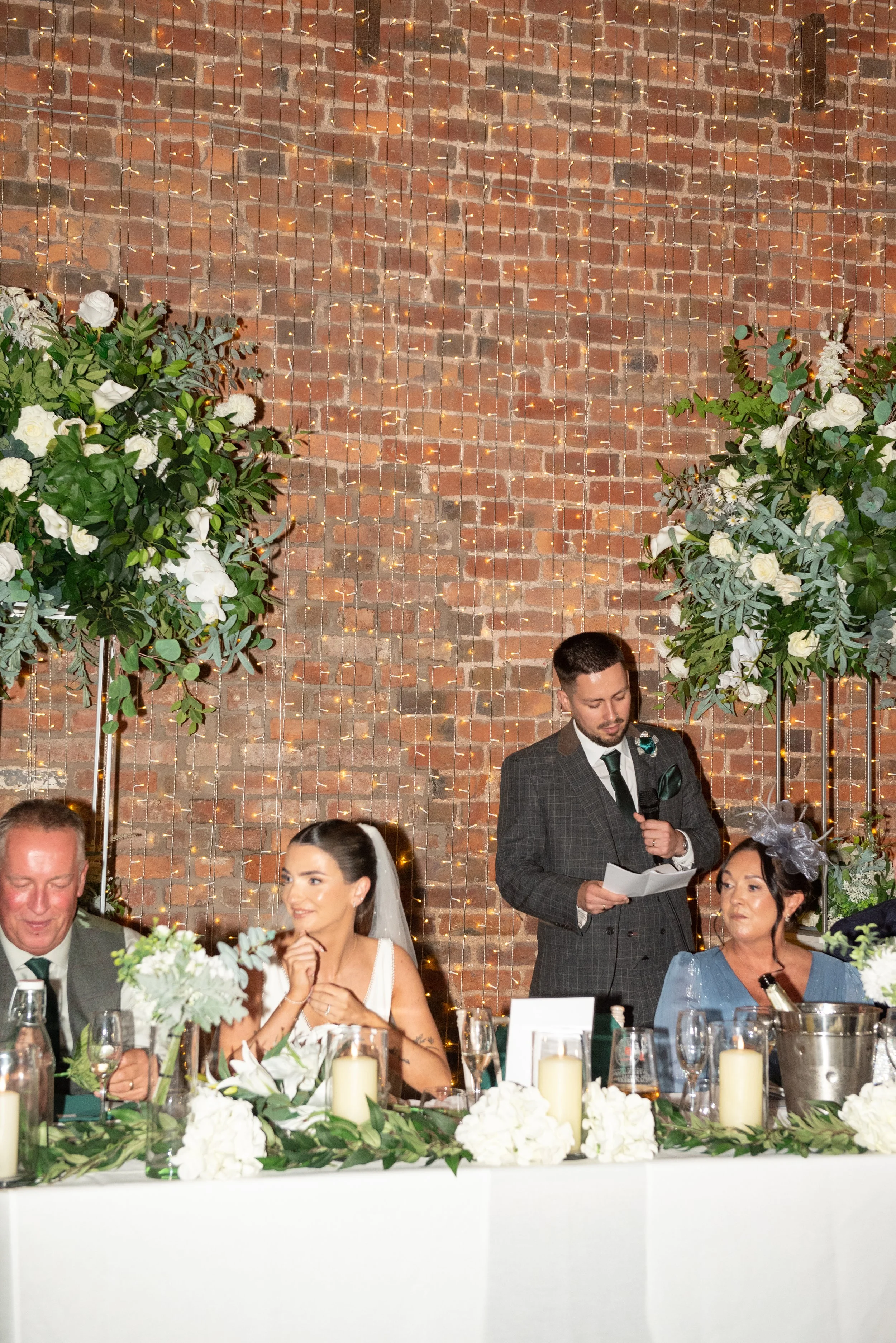 A wedding reception with a bride, a groom, and two women seated at a decorated table, with a man giving a speech or toast.