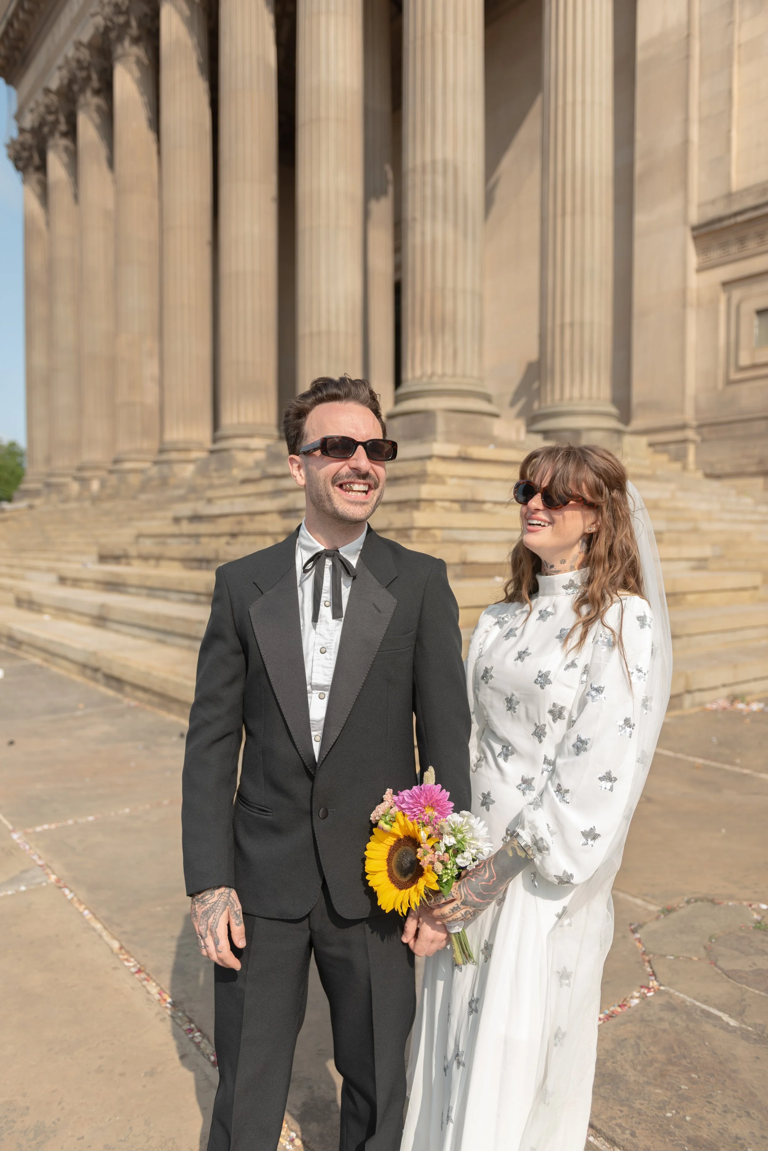 A bride and groom stand outside a historic building with large columns, holding hands, smiling, wearing sunglasses, the bride holding a bouquet of flowers, including sunflowers, on a sunny day.