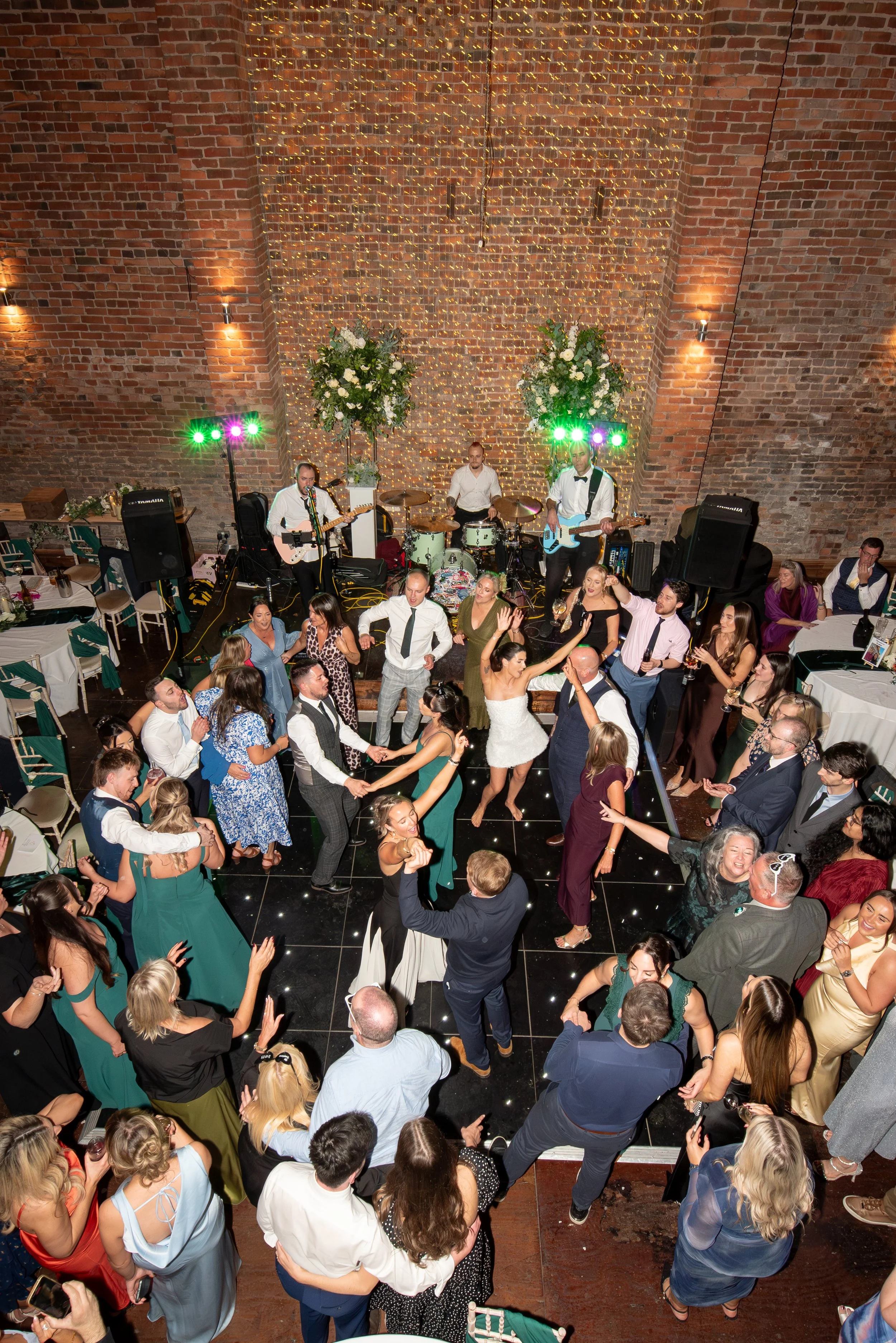 People dancing and enjoying live music at a wedding reception in a rustic brick venue with string lights and floral arrangements.