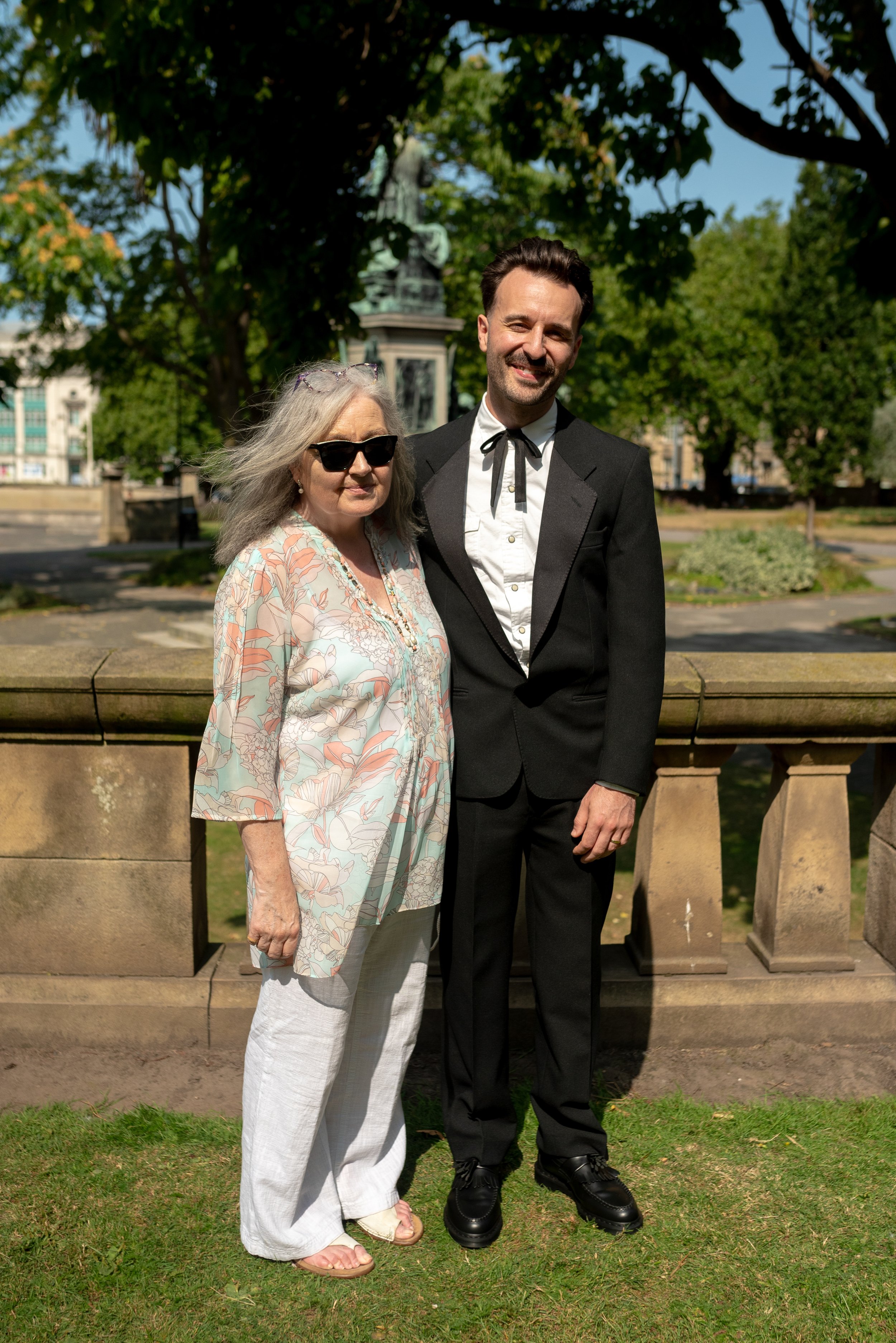 A woman in sunglasses and a floral blouse standing beside a man in a tuxedo in a park, with a statue and trees in the background.