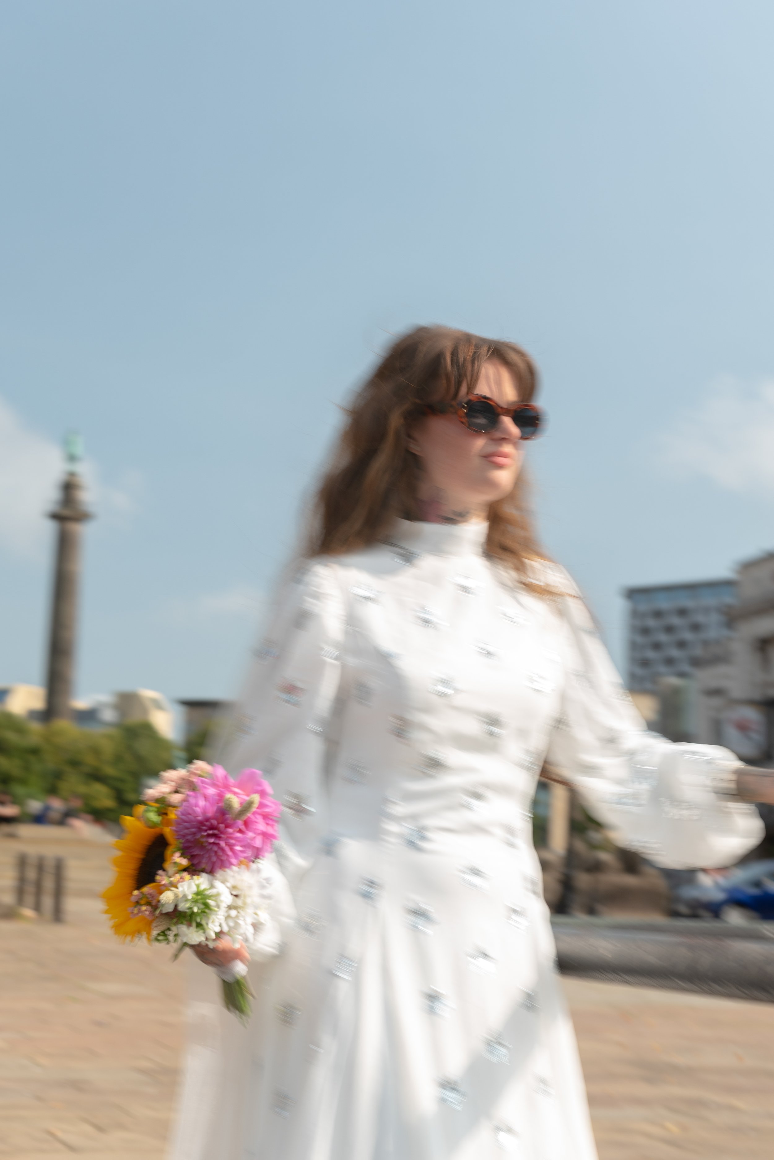 A woman in a white dress holding a colorful bouquet of flowers, walking outdoors with a cityscape background.